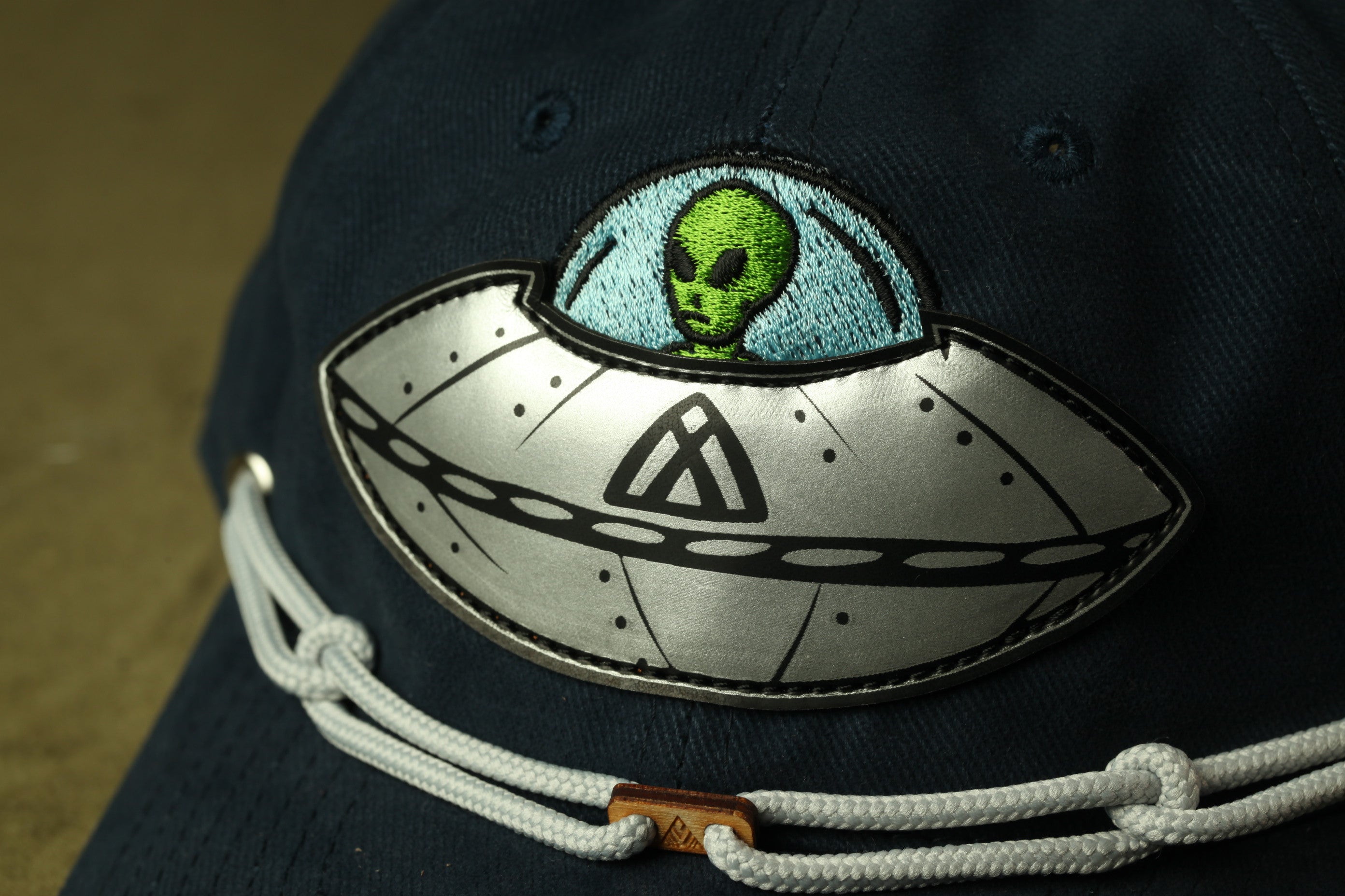 a close-up view of a dark blue baseball cap with a unique embroidered design on the front. The design includes a green alien character inside a silver spaceship, which is a distinctive and eye-catching feature of the cap.