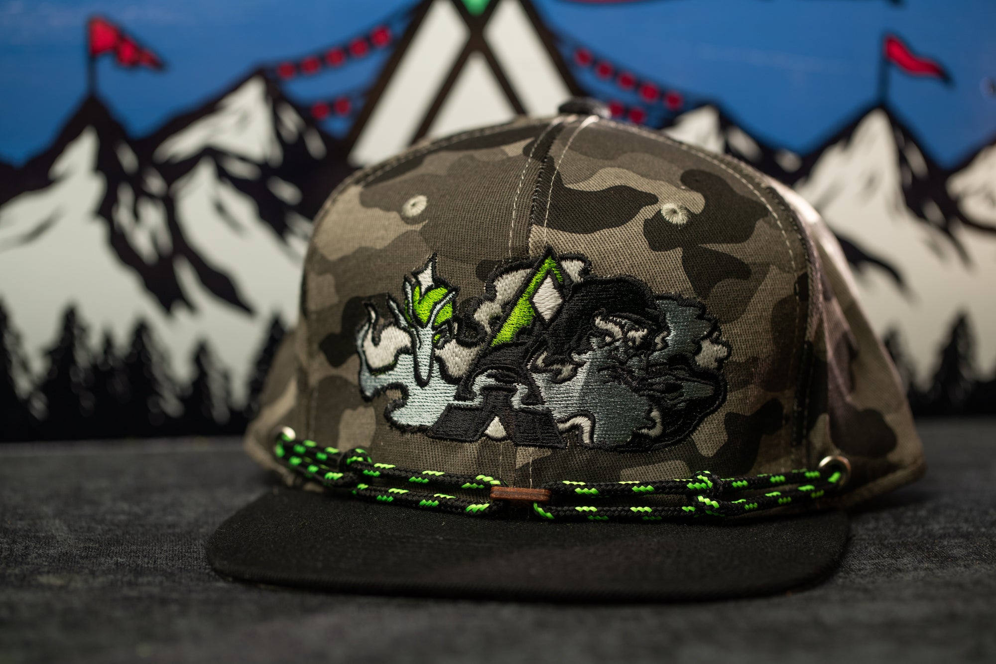 A camouflage baseball cap with a graphic design on the front, placed on a surface with a mountain landscape in the background.