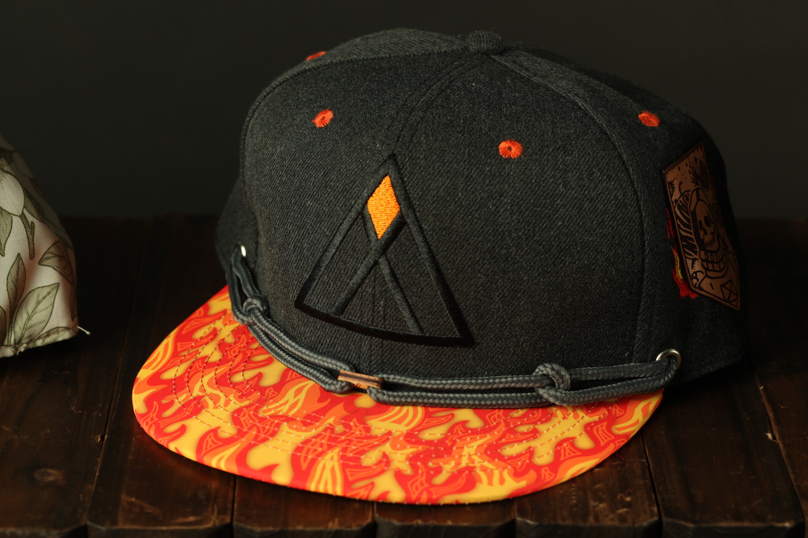 A black baseball cap with a yellow and orange flame pattern on the brim and a triangular logo on the front, placed on a wooden surface.