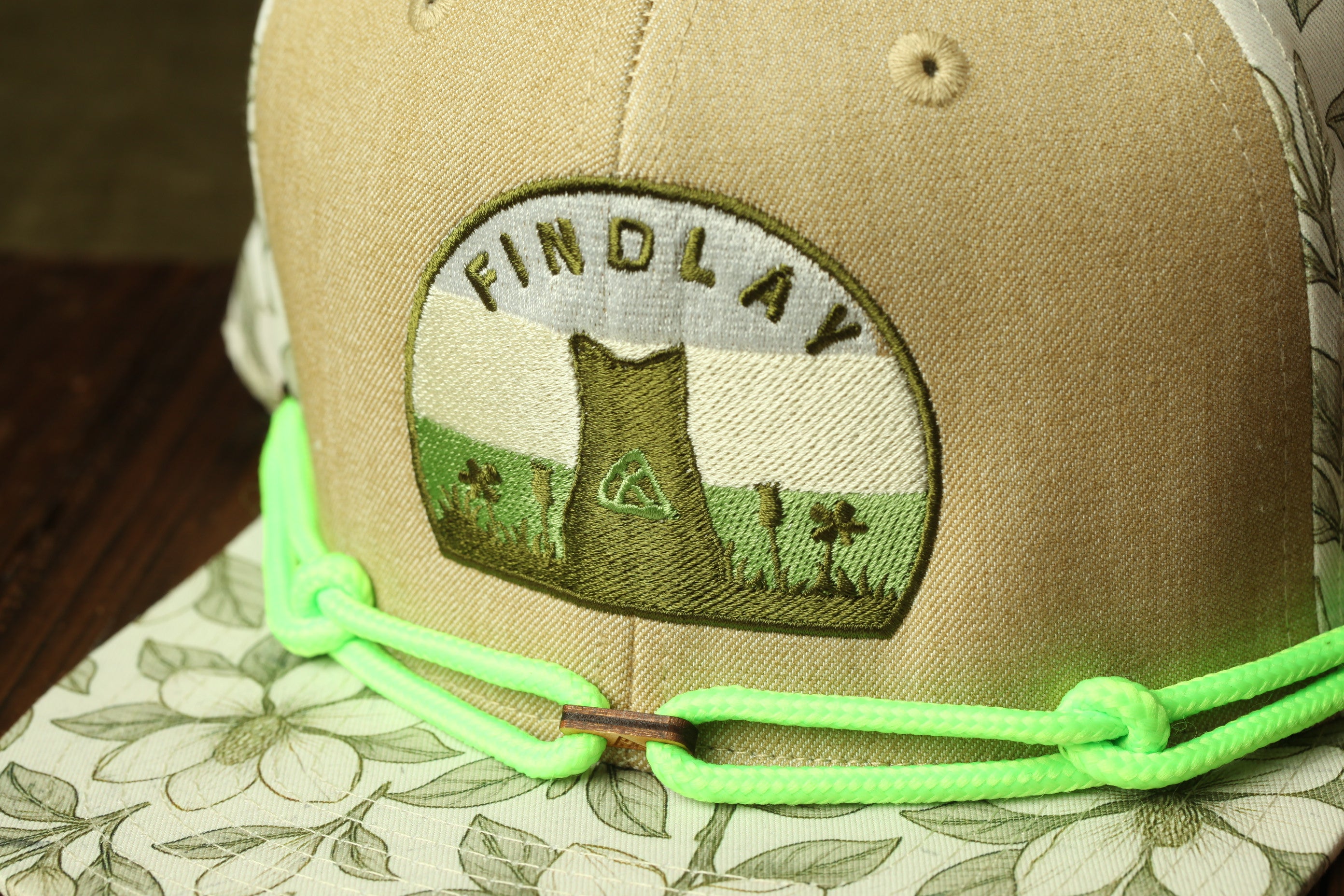 A close-up view of a beige baseball cap with a green string and a patch on the front that says "FINDLAY".