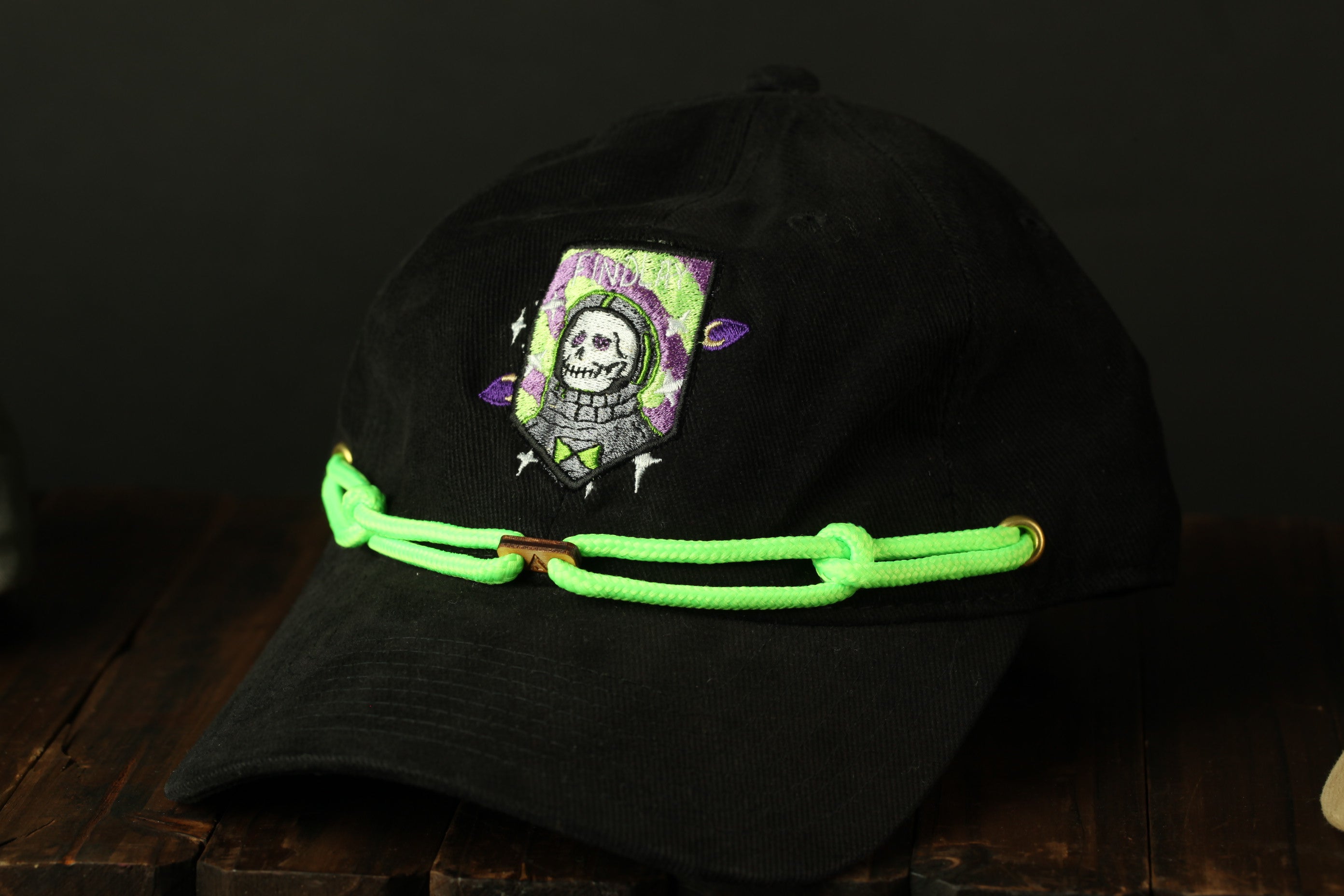 A black baseball cap with a skull and crossbones design on the front, and neon green strings around the brim.