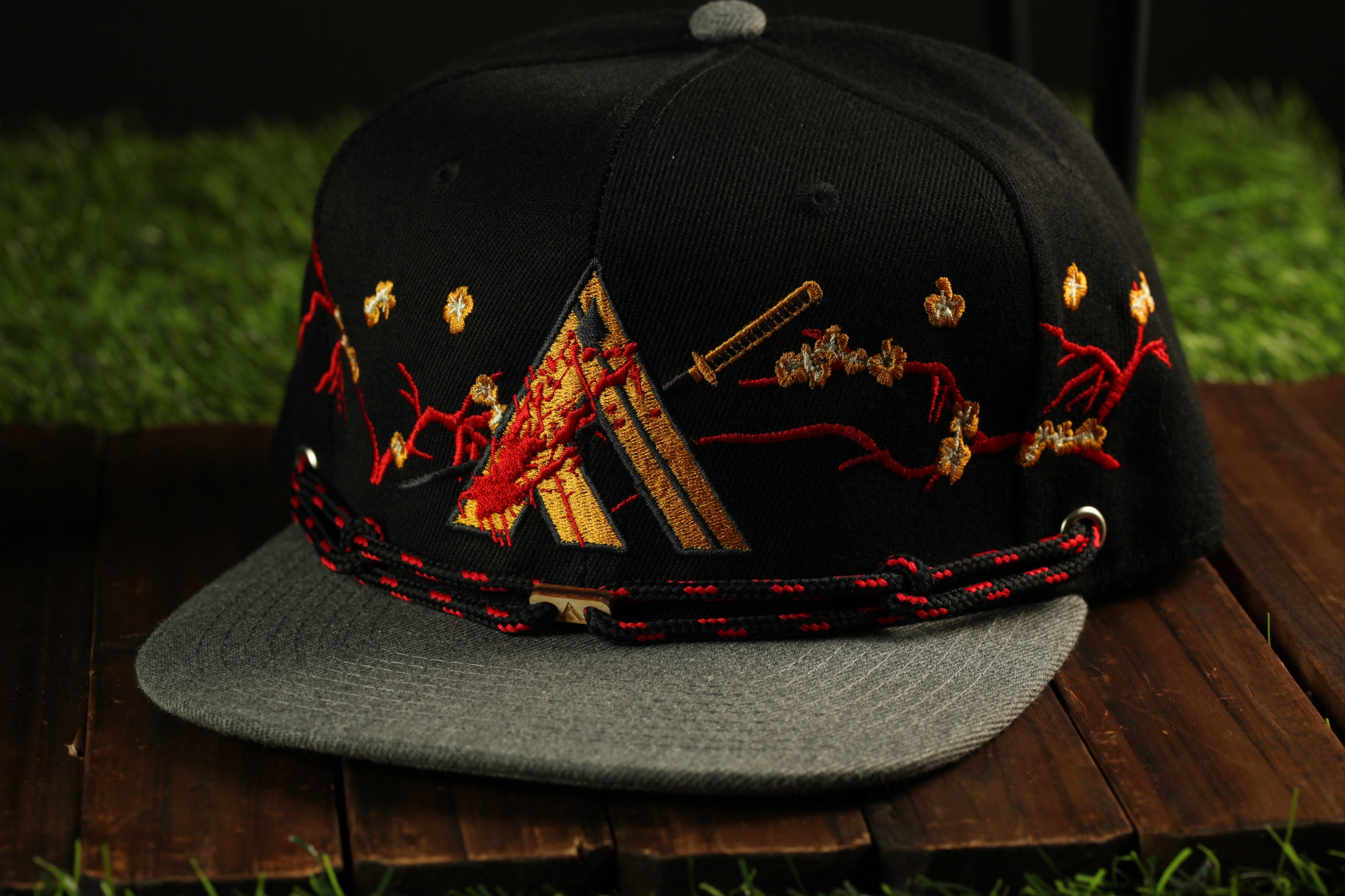 A black baseball cap with gold and red embroidered designs, including a triangle and floral patterns, is displayed on a wooden surface.