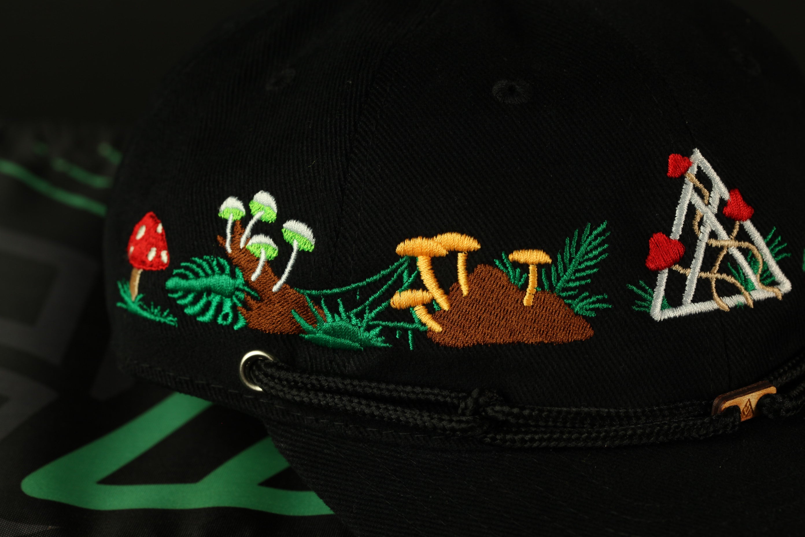 a close-up view of a black baseball cap with colorful embroidered designs, including mushrooms, leaves, and other natural elements.