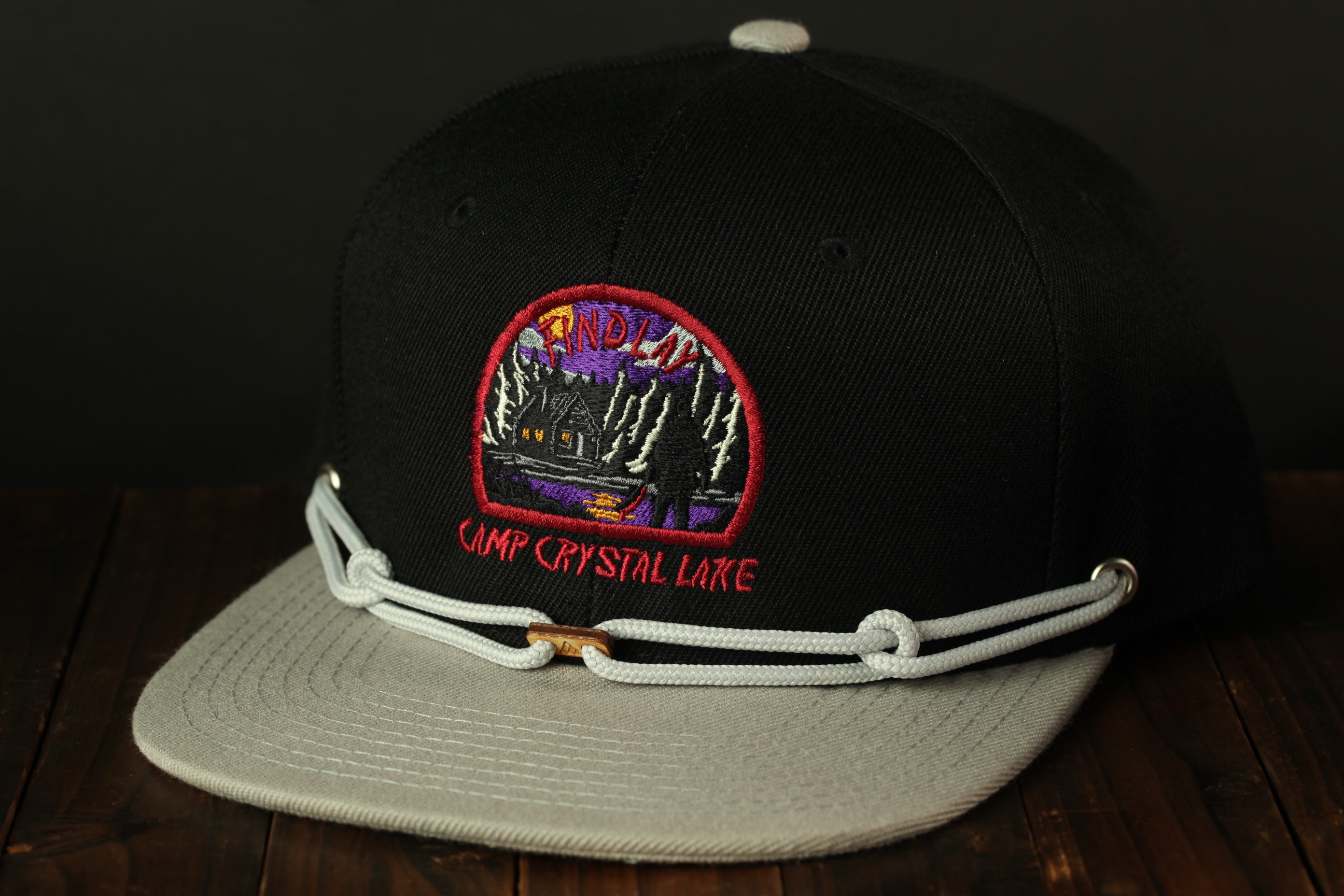 A black baseball cap with a white brim and a colorful embroidered patch on the front, featuring a scenic landscape with a cabin and trees.