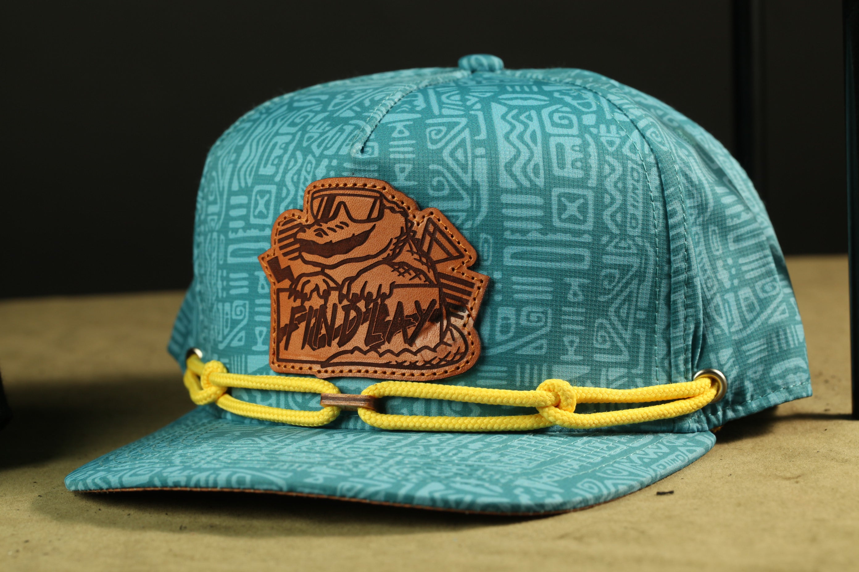A turquoise baseball cap with a leather patch featuring a cartoon character and yellow straps.