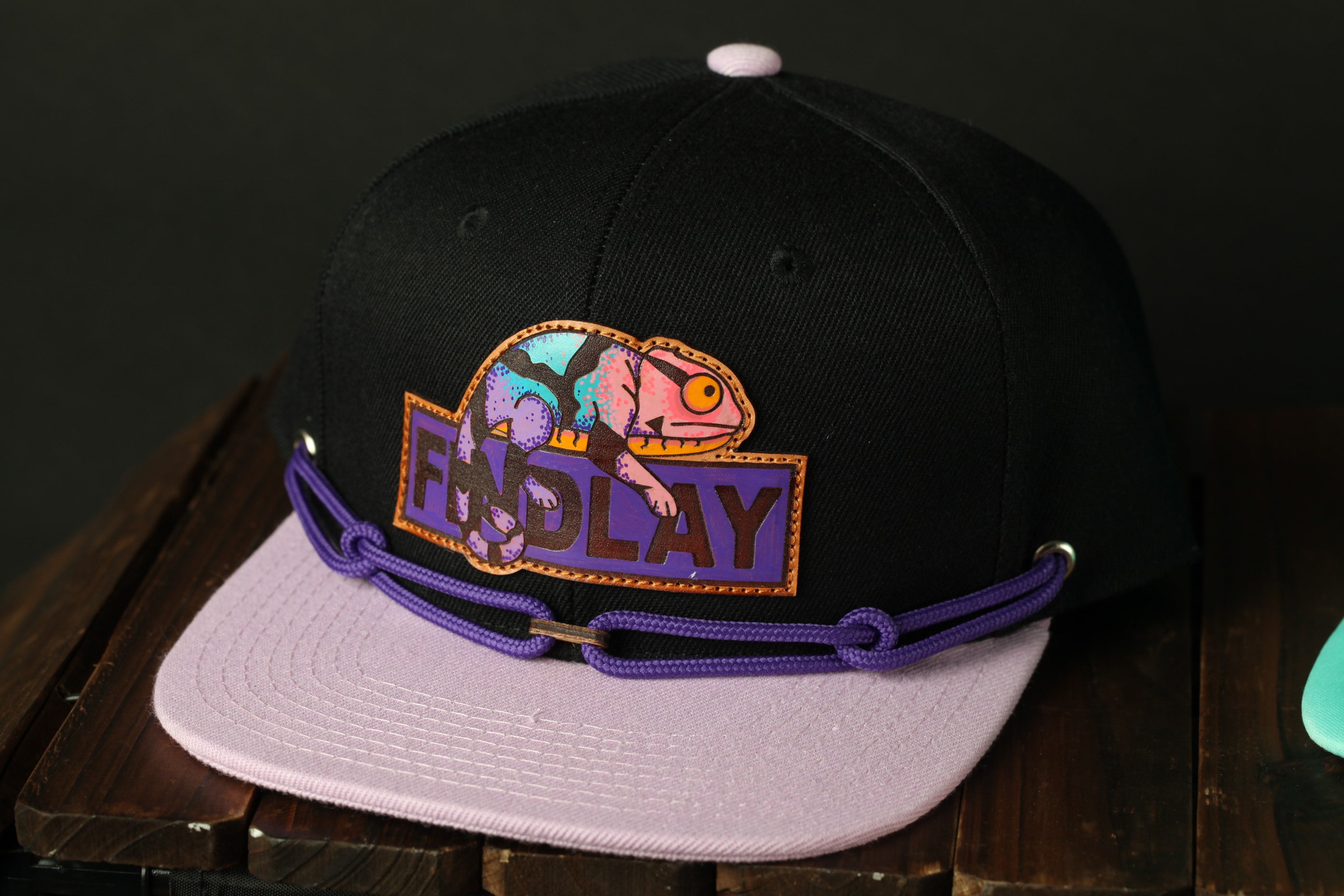A black baseball cap with a colorful graphic and the word "FINDlay" printed on it, sitting on a wooden surface.
