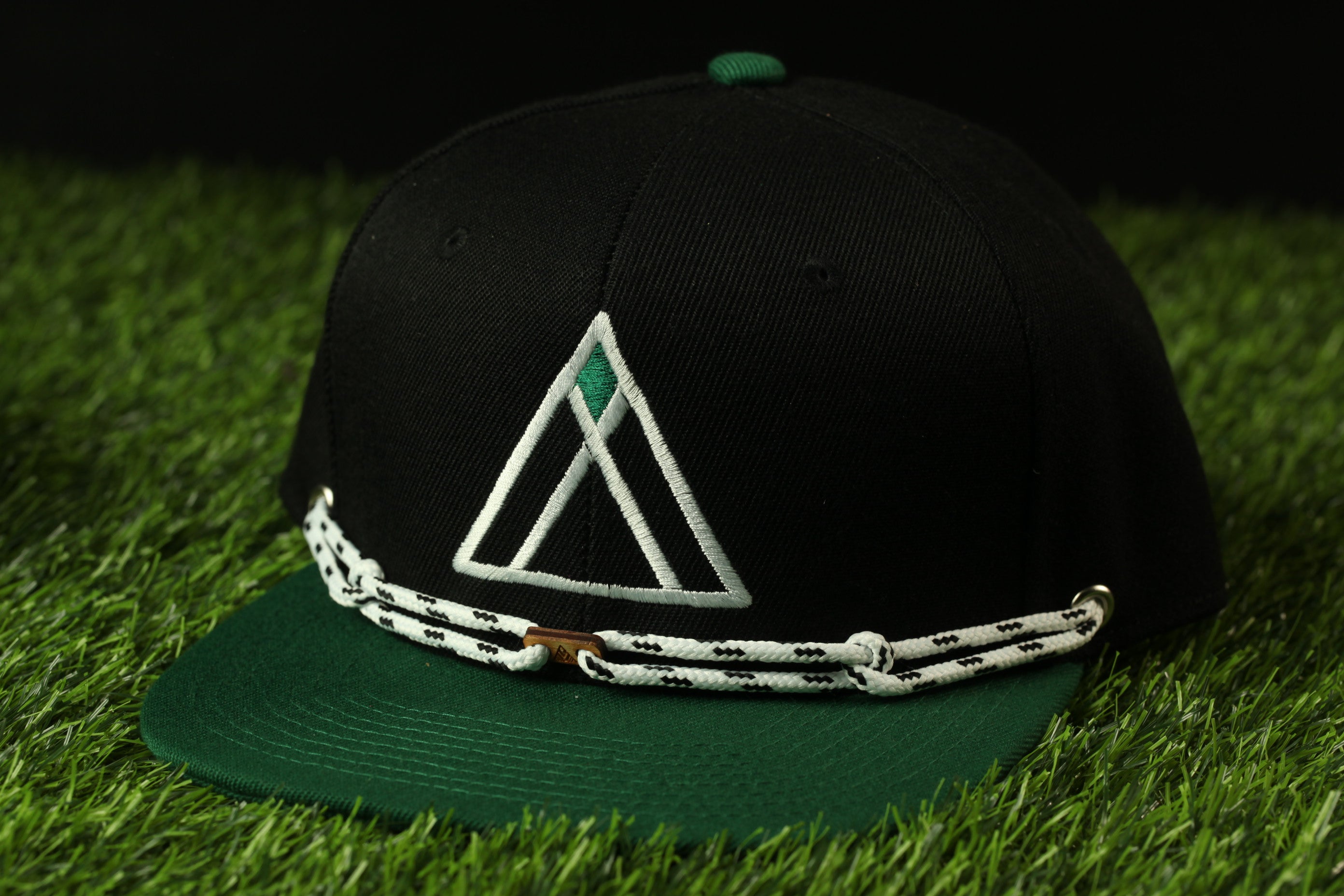 A black baseball cap with a green brim and a white triangular logo on the front, resting on a grassy surface.