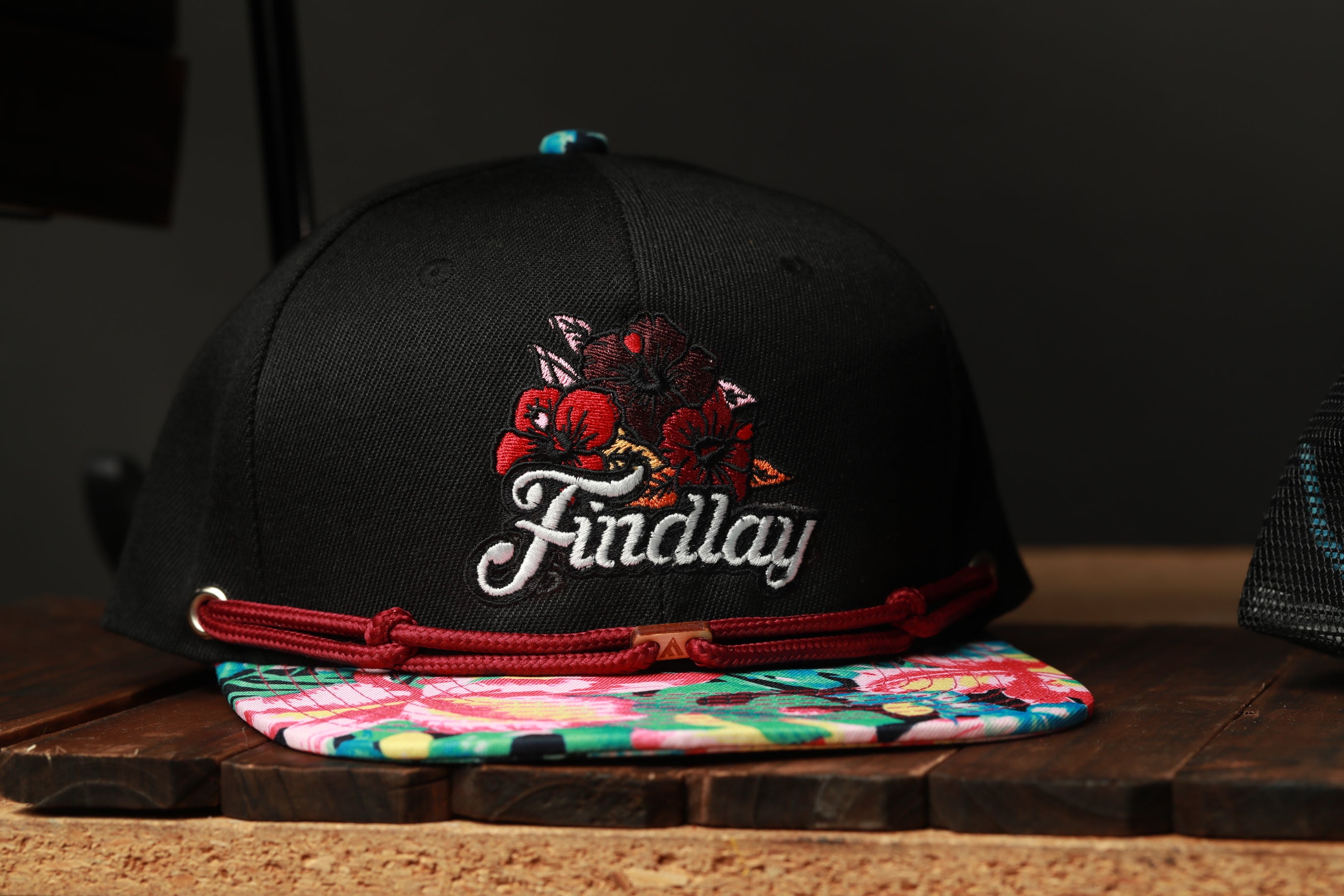 A black baseball cap with a floral embroidered logo and a colorful printed cap lying on a wooden surface.