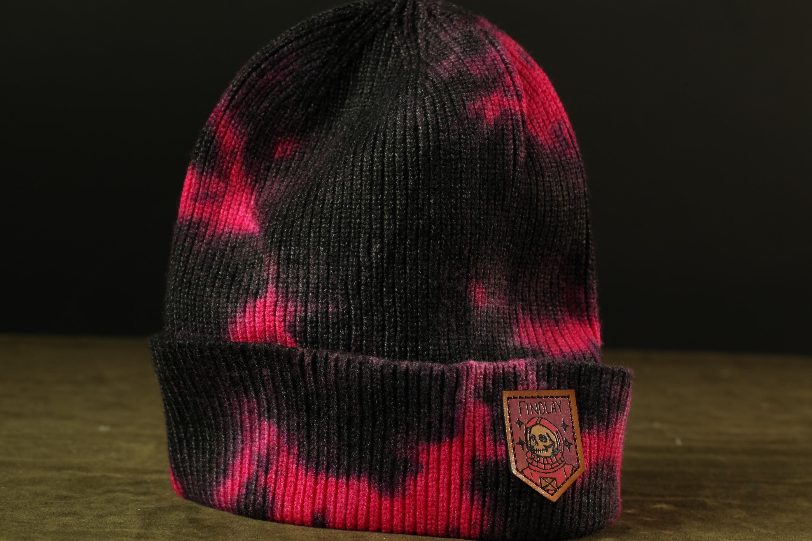 A black and pink knitted beanie with a patch on the front, featuring a skull design.