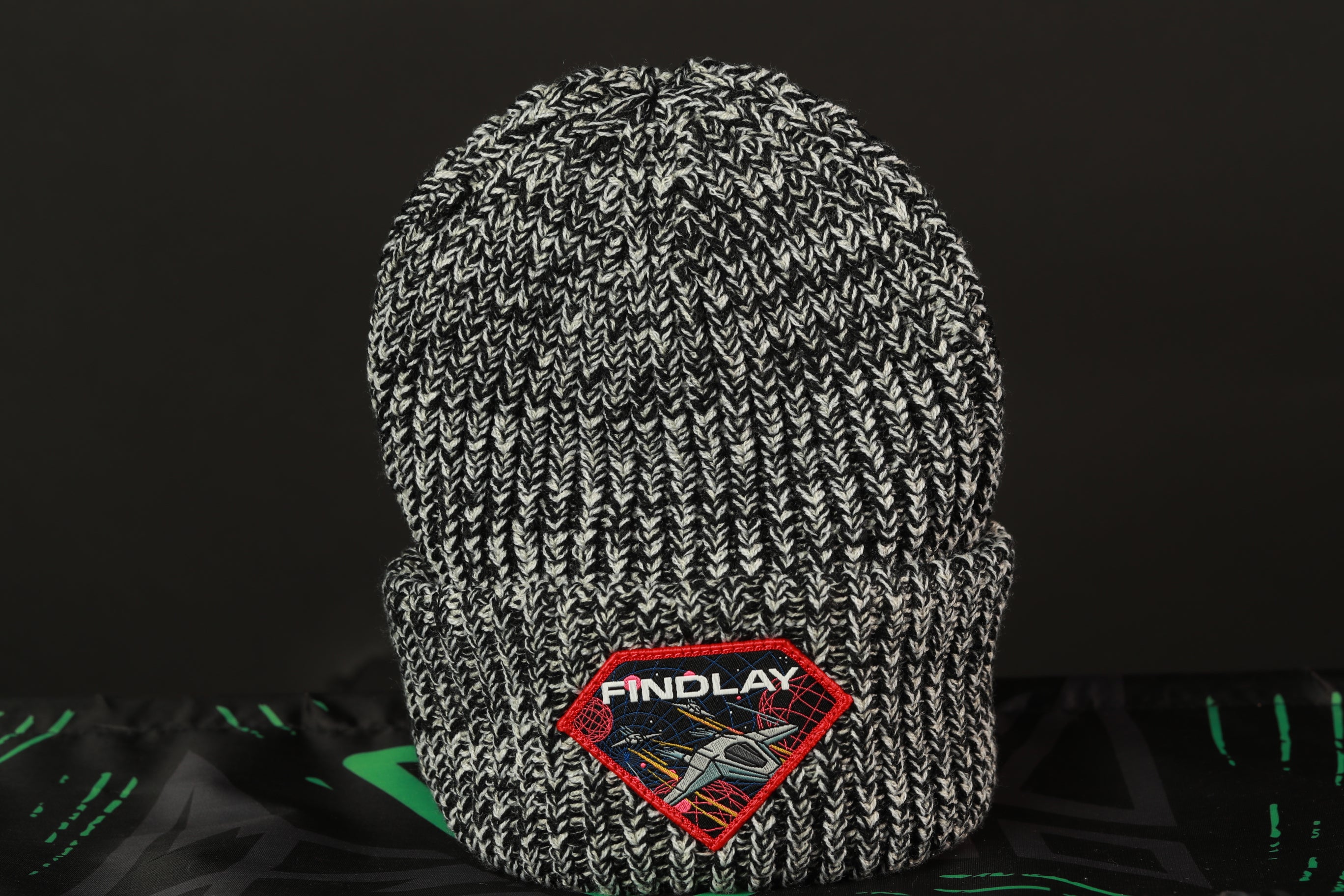 A black and white knit beanie with a red and white patch on the front, featuring the text "FINDLAY" and an illustration of a rocket ship.
