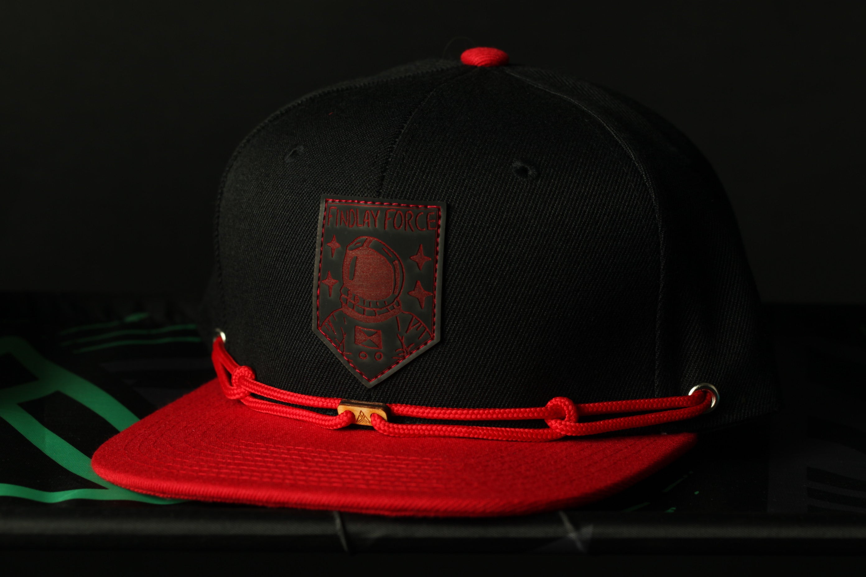 A black baseball cap with a red brim and a red and black patch on the front, featuring a graphic design.