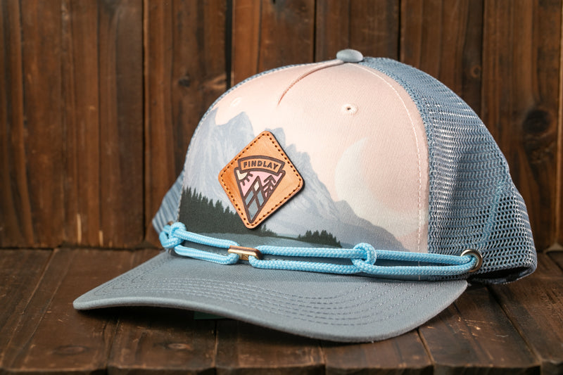 A blue and gray baseball cap with a mountain landscape design and a leather patch on the front, resting on a wooden surface.