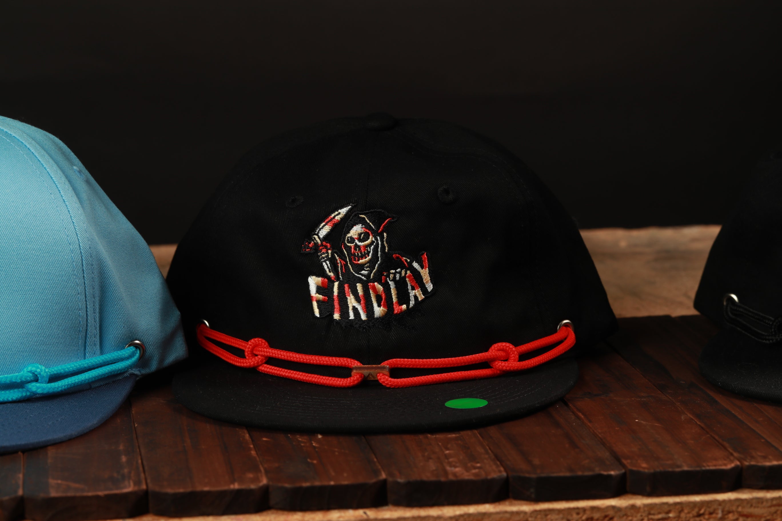 A black baseball cap with a red chain strap and a graphic of a skull wearing a hood, sitting on a wooden surface.