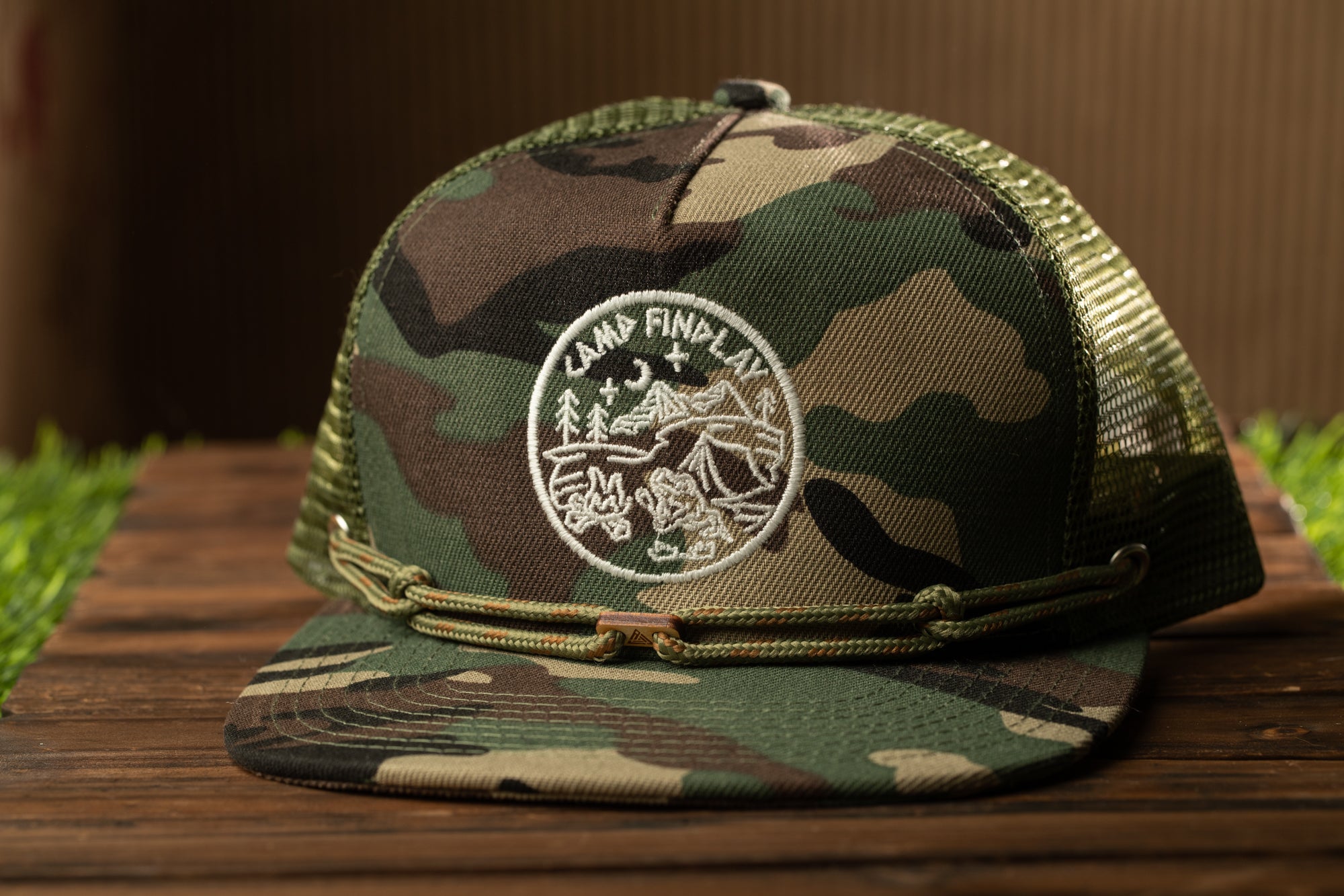 A camouflage baseball cap with a logo on it, placed on a wooden surface.