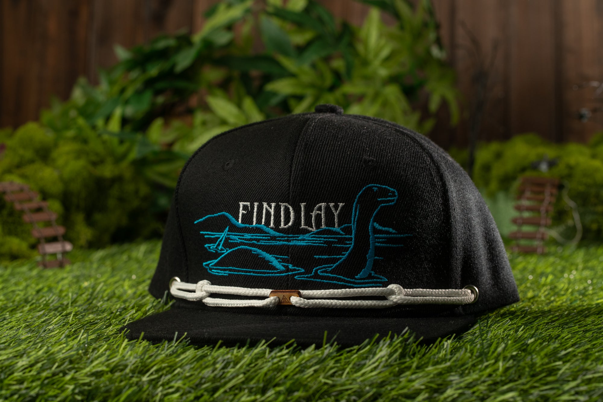 A black baseball cap with a blue and white design, featuring a cartoon-like creature, is placed on a grassy surface with a wooden fence and greenery in the background.