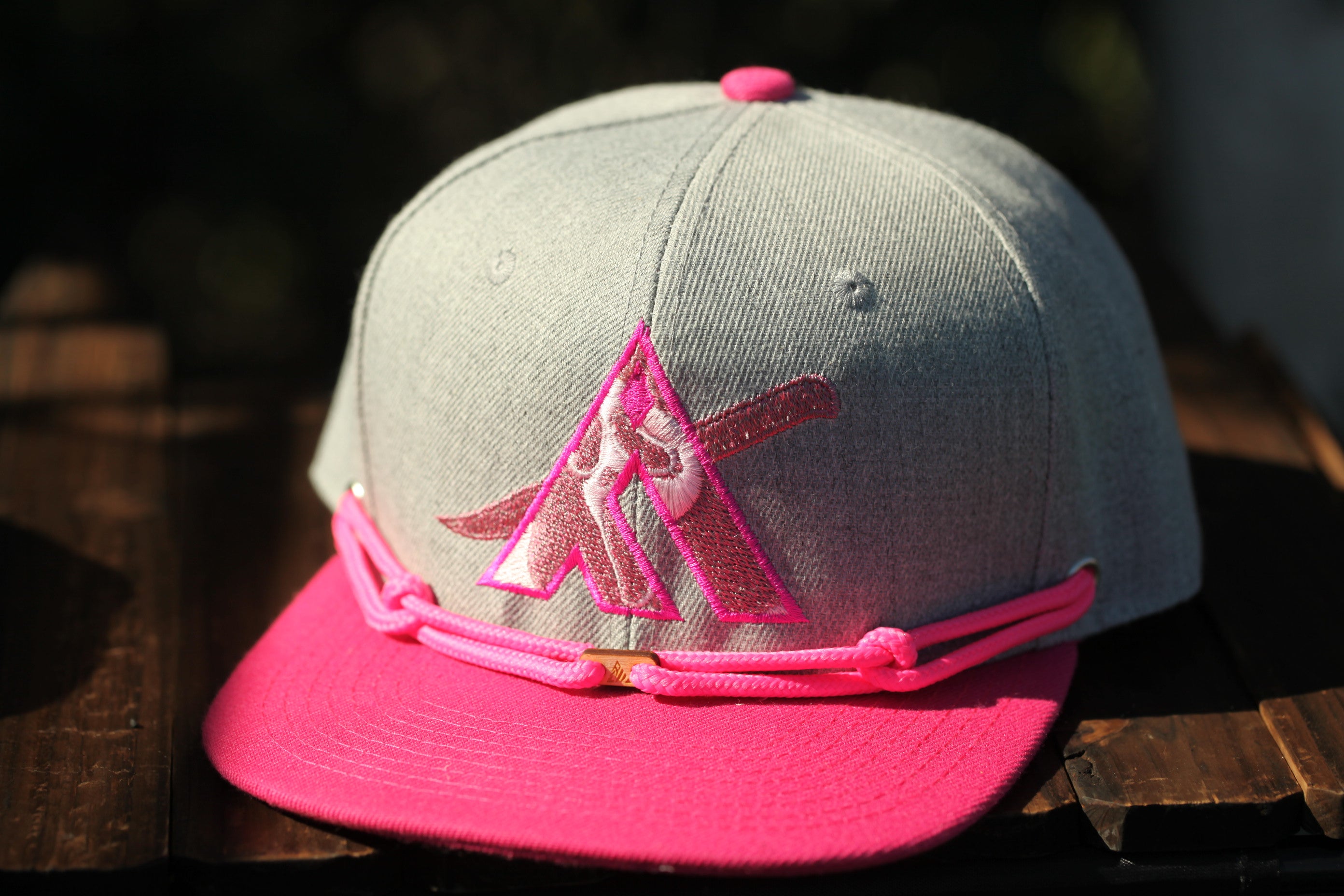 A gray baseball cap with a pink brim and a pink logo on the front, placed on a wooden surface.