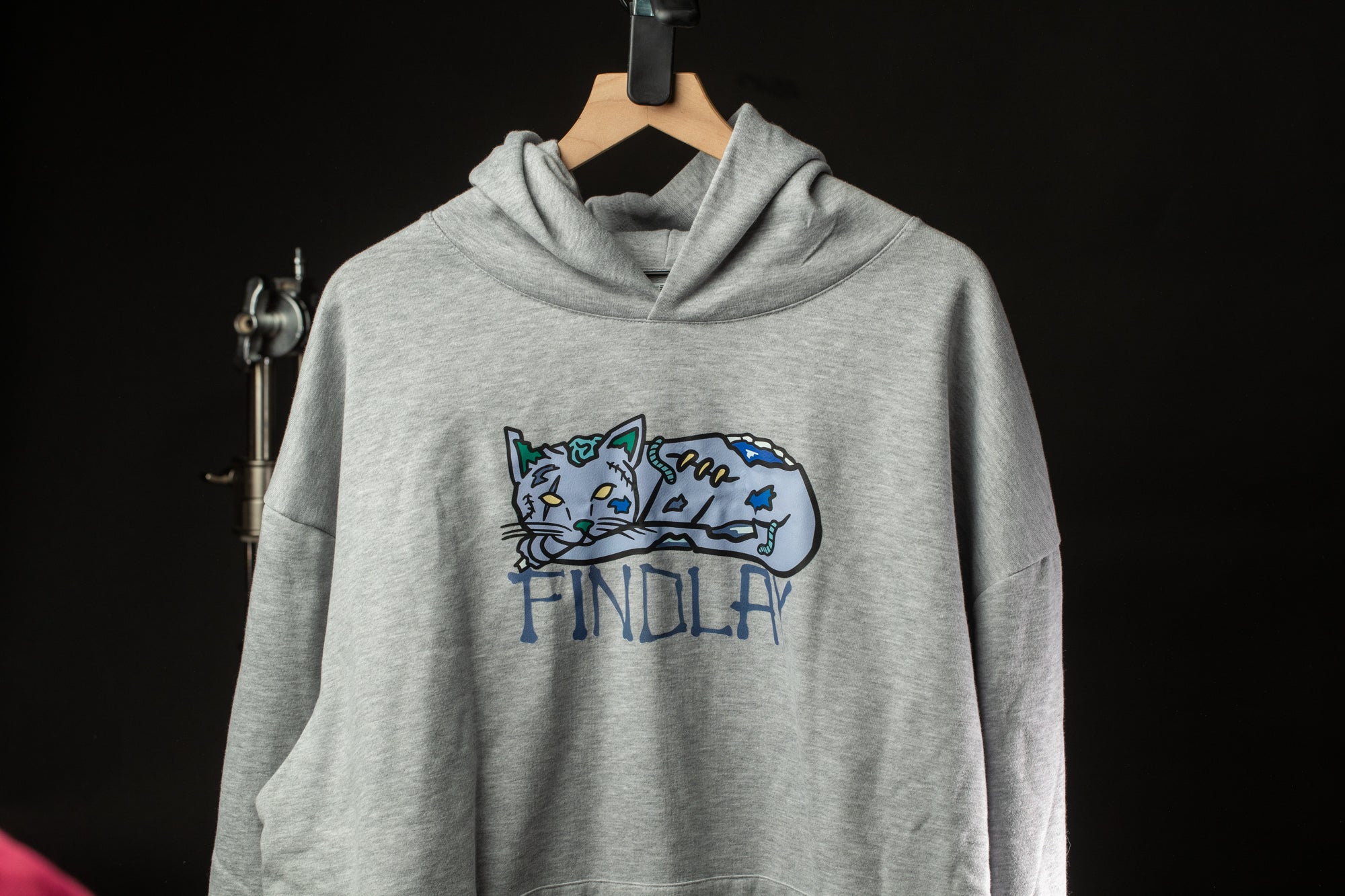 A grey hoodie with a cat design and the word "FINDOLAY" printed on it, hanging on a wooden hanger against a black background.