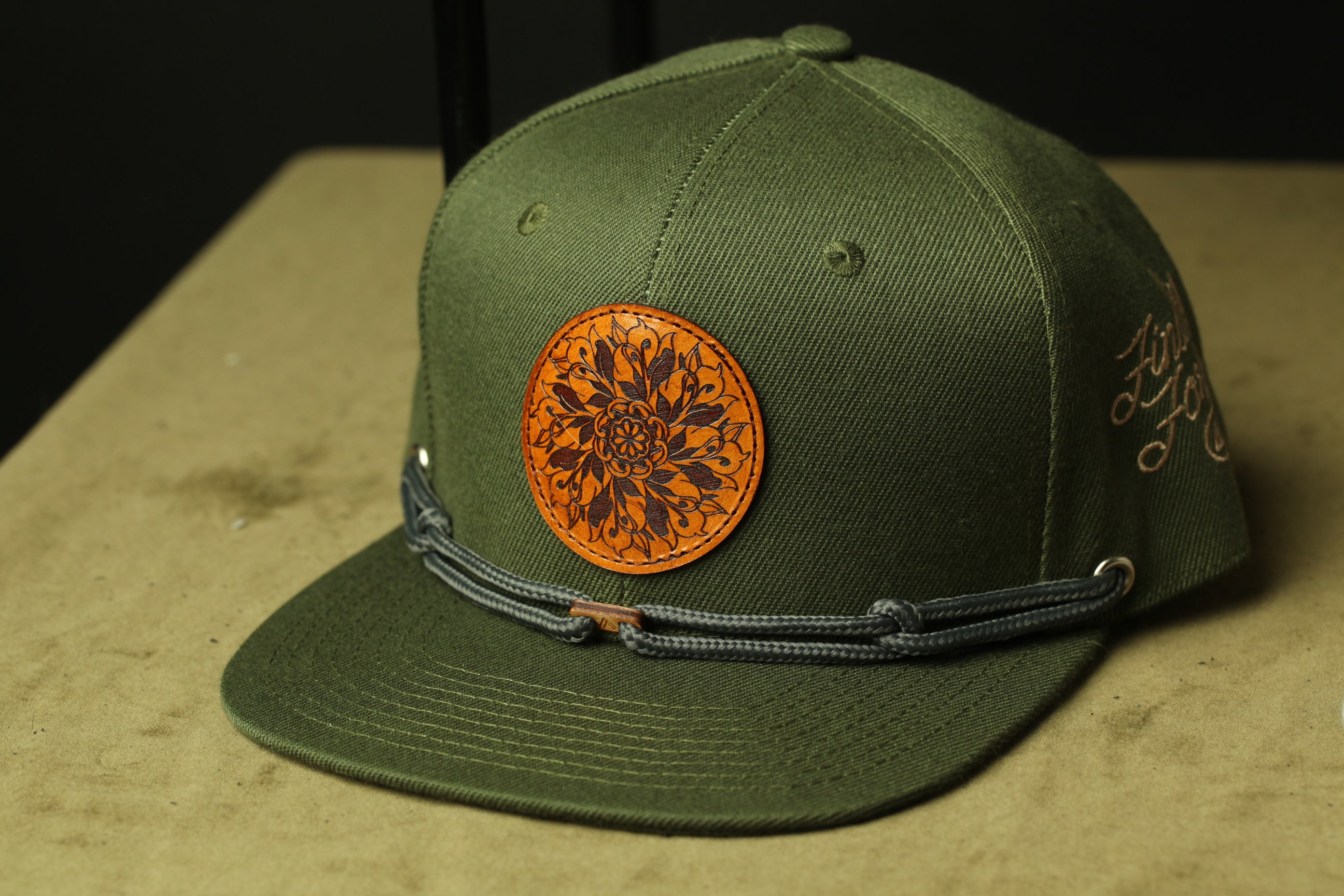 A green baseball cap with a circular leather patch featuring an intricate floral design.