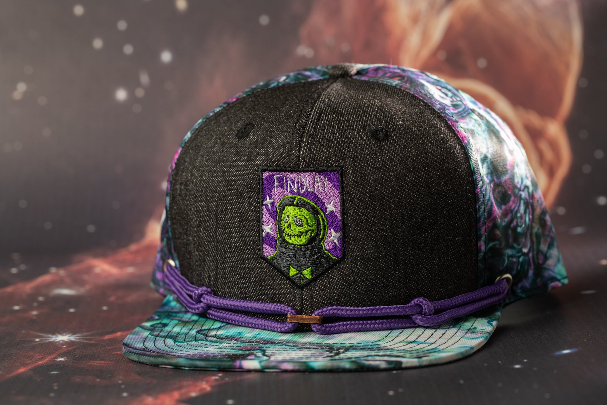 A black baseball cap with a purple and green patterned brim, featuring a patch with a skull and crossbones design.