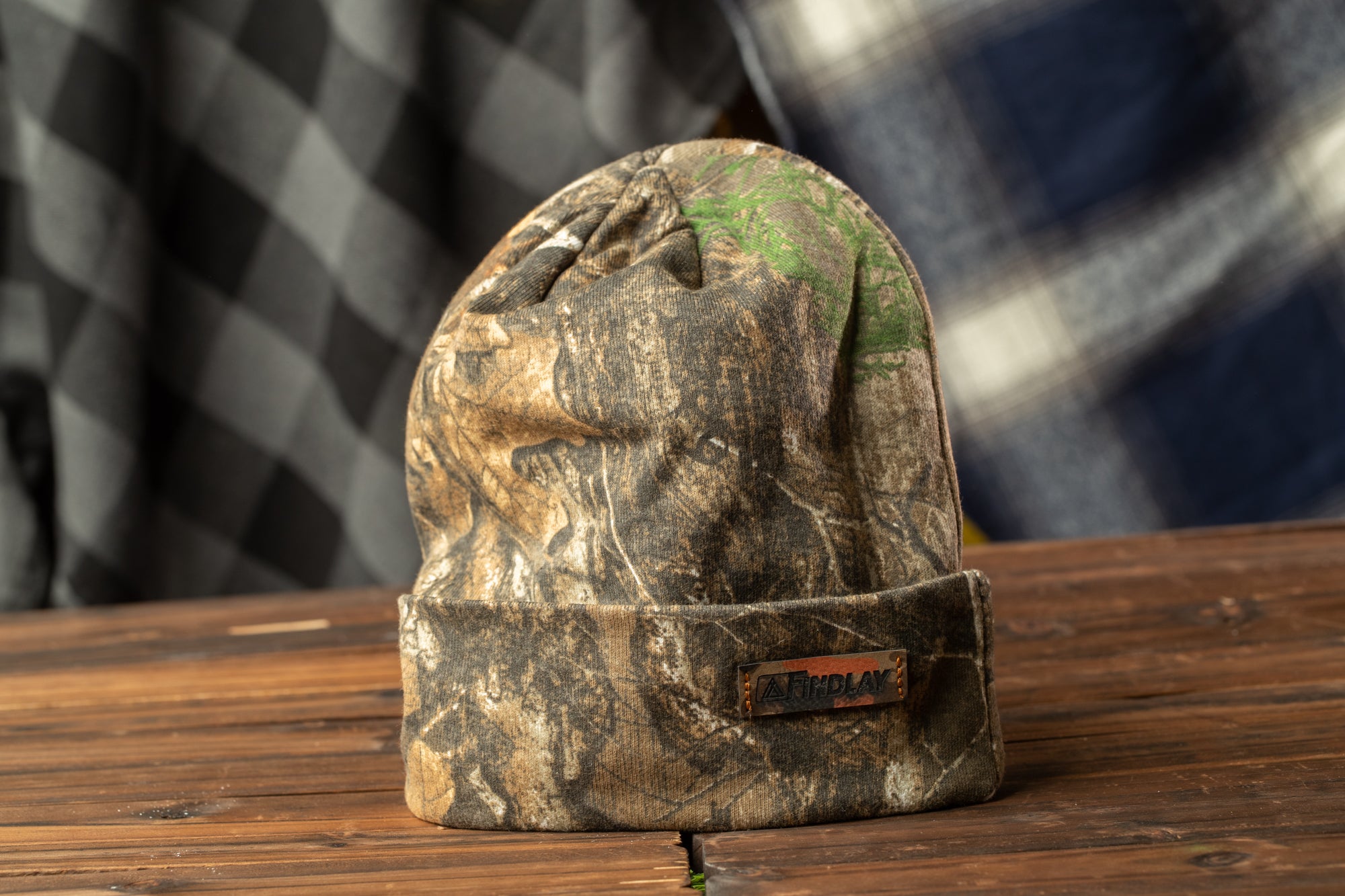 A camouflage-patterned beanie with a label on it, placed on a wooden surface against a checkered background.