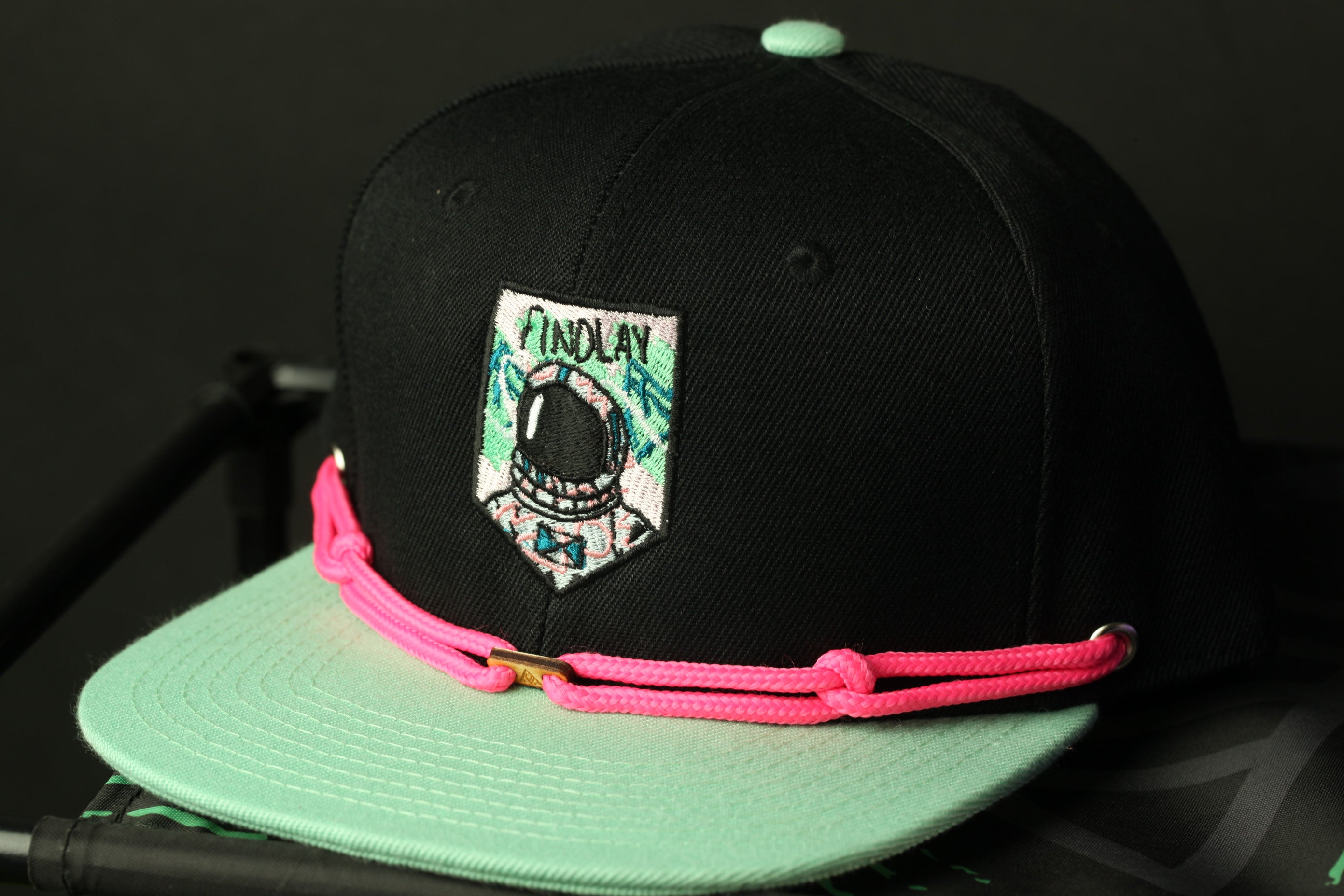A black baseball cap with a pink and green strap, featuring a patch with a graphic of an astronaut and the word "FINDLAY" on it.