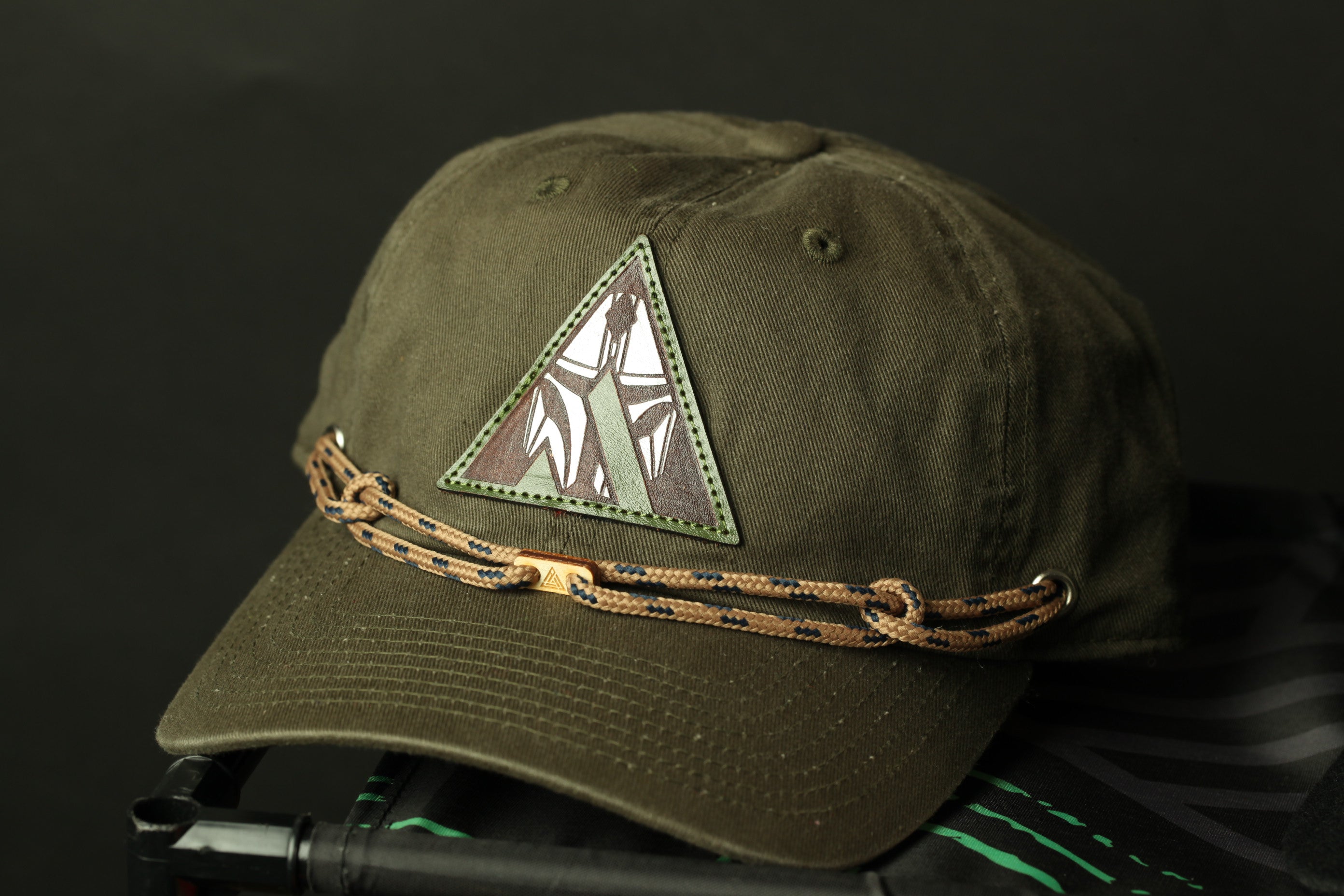 A dark green baseball cap with a triangular patch on the front, featuring a design, is shown against a dark background.
