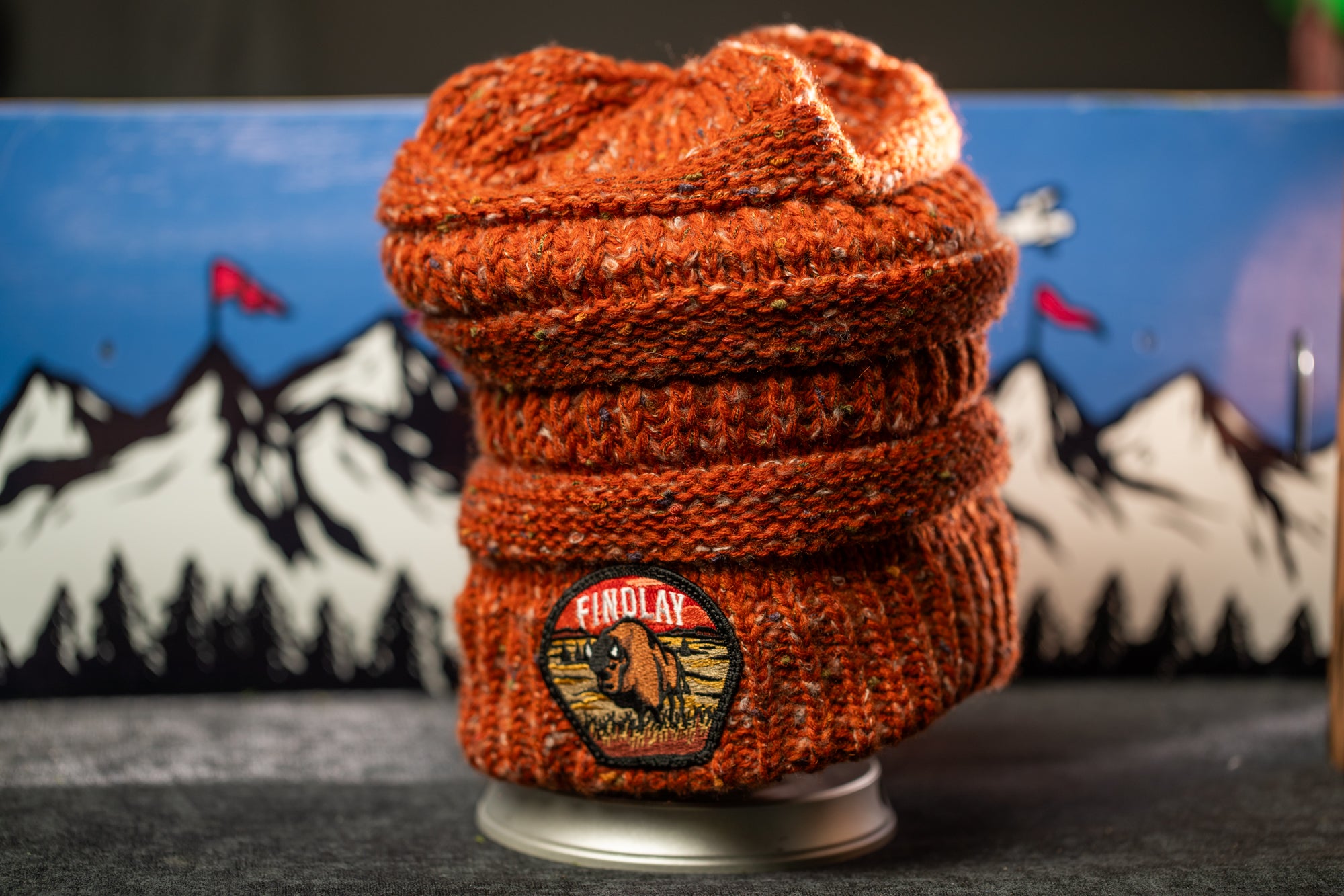 An orange knit beanie with a patch on the front, placed on a gray surface against a backdrop of mountains and flags.