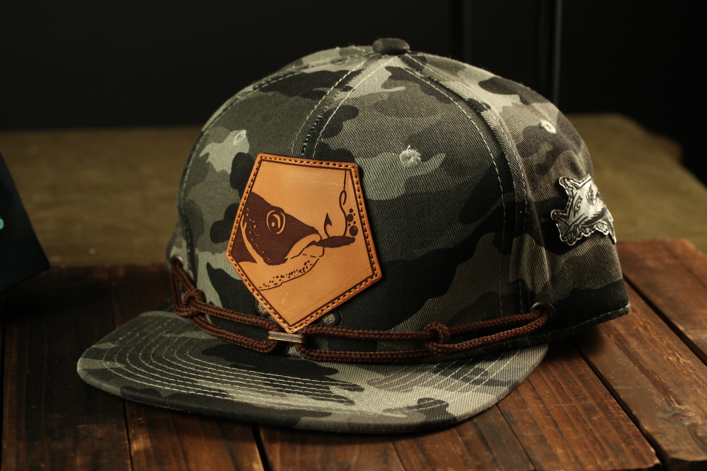 A camouflage baseball cap with a leather patch featuring a fish design, placed on a wooden surface.