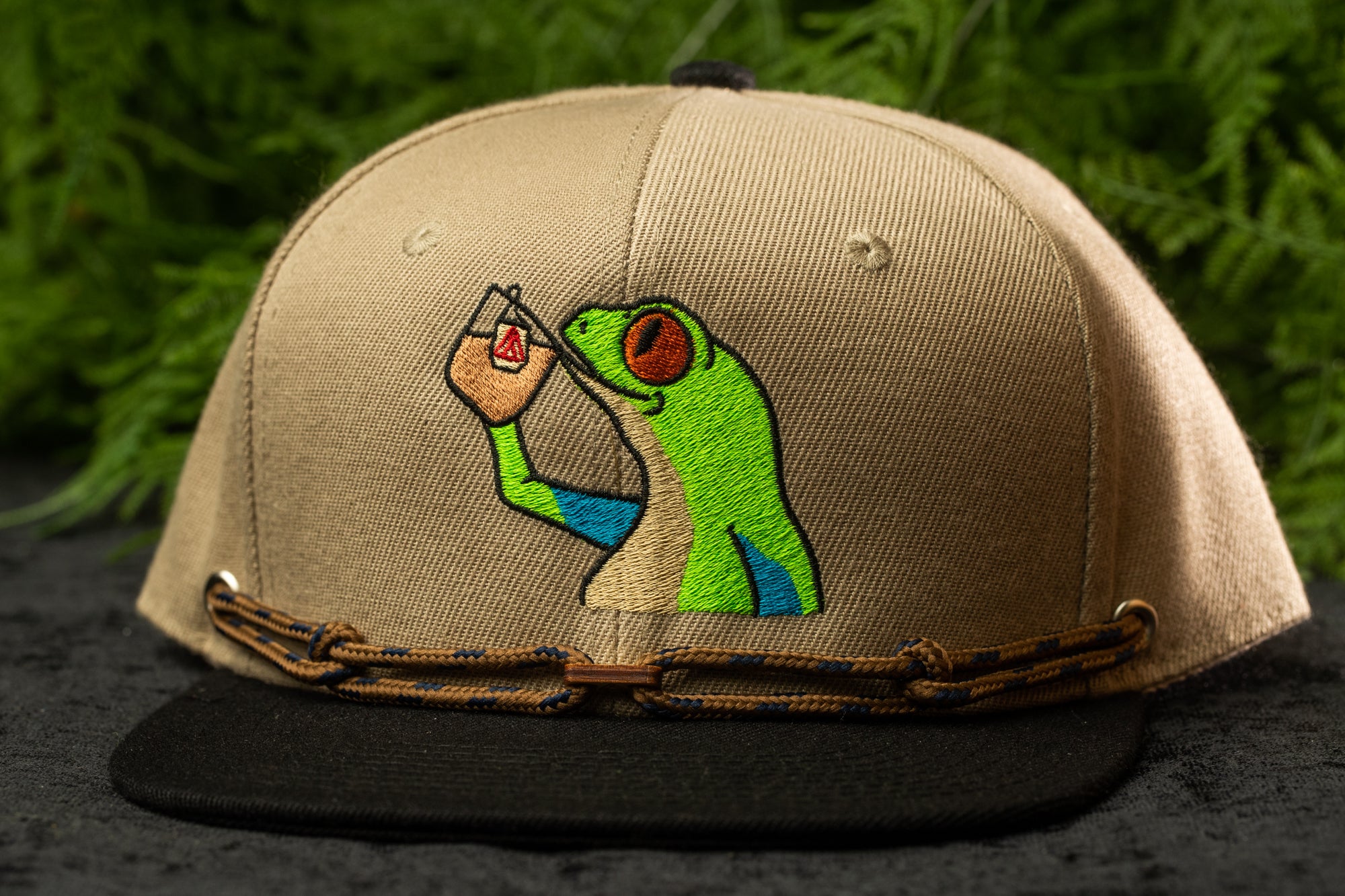 A beige baseball cap with a green frog embroidered on it, holding a drink in its hand.