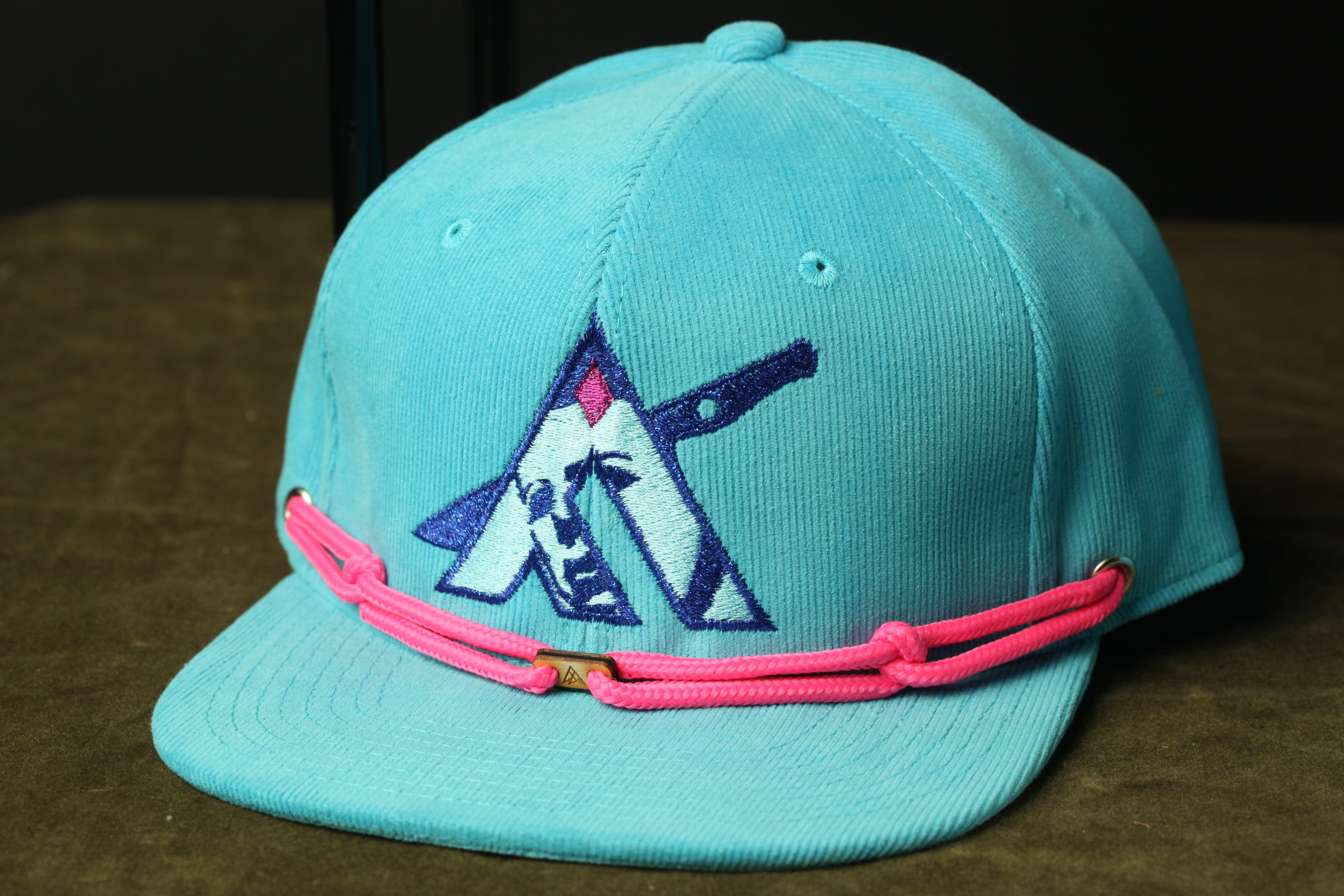 A turquoise baseball cap with a pink braided strap and a logo on the front.