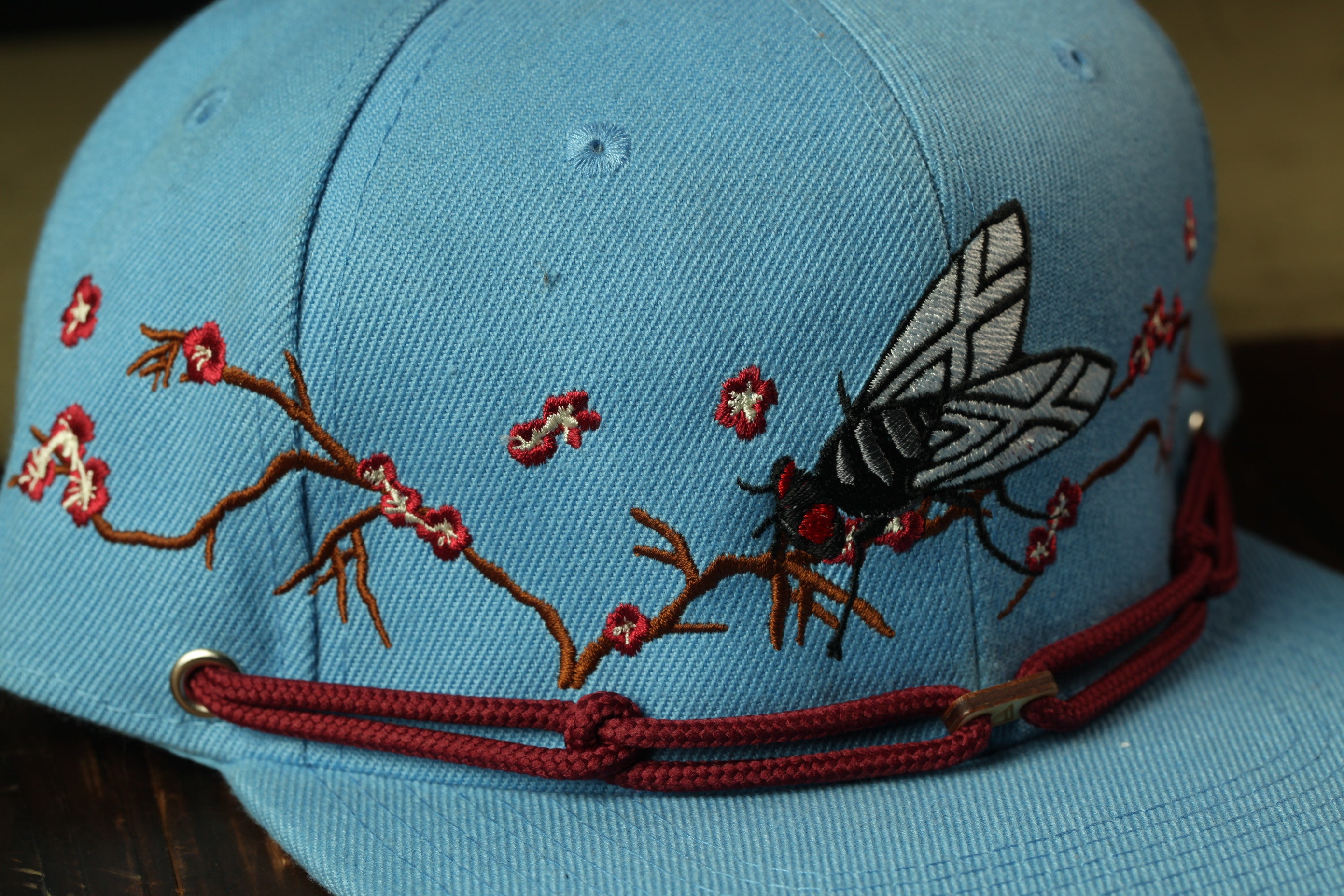 A close-up view of a blue baseball cap with a detailed embroidered design of a fly and flowers on the brim.