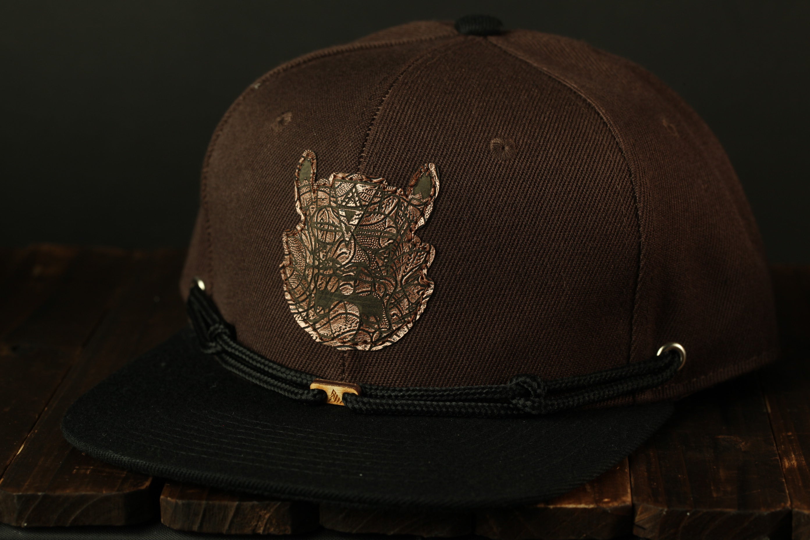 A brown baseball cap with a metal emblem on the front, sitting on a wooden surface.