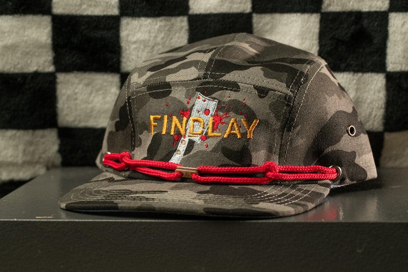 A camouflage baseball cap with the word "FINDOLAY" embroidered on it, sitting on a black surface against a checkered background.