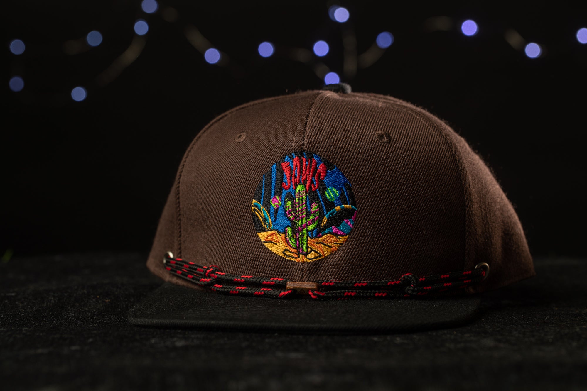 A brown baseball cap with a colorful embroidered design on the front, featuring a cactus and other desert-themed elements.