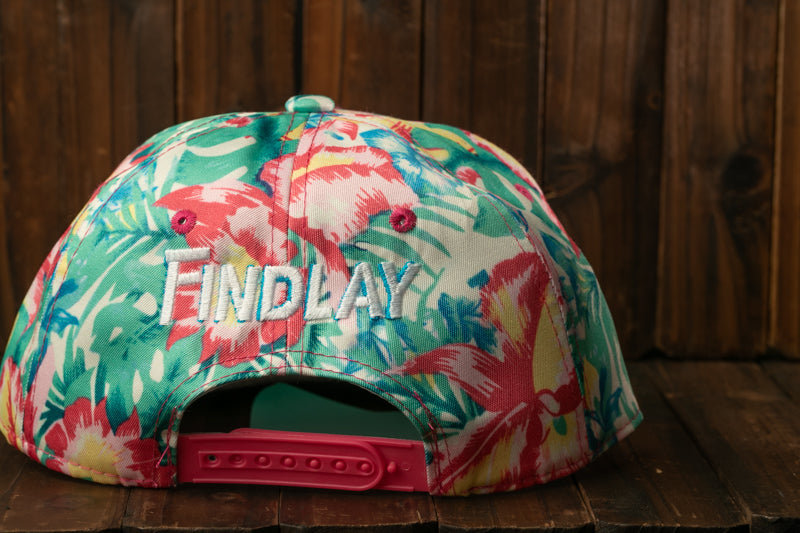A colorful floral print baseball cap with the word "Findlay" embroidered on it, sitting on a wooden surface.