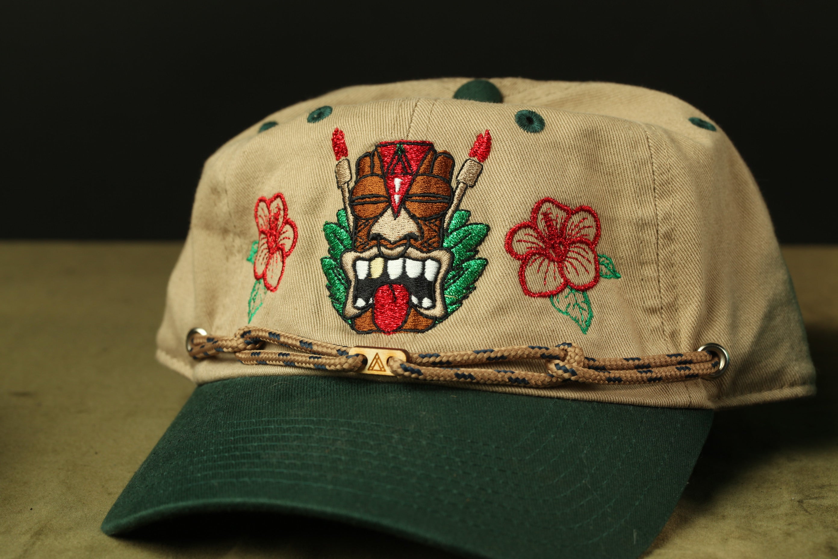 A beige baseball cap with a tiki mask design and red flowers on the brim.
