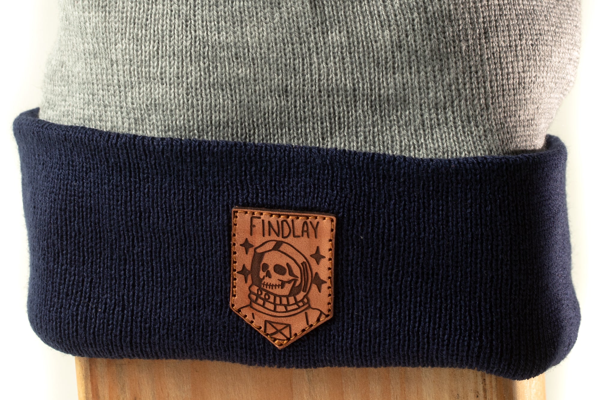 A close-up view of a dark blue knit beanie with a navy blue trim, featuring a leather patch with a skull and crossbones design.