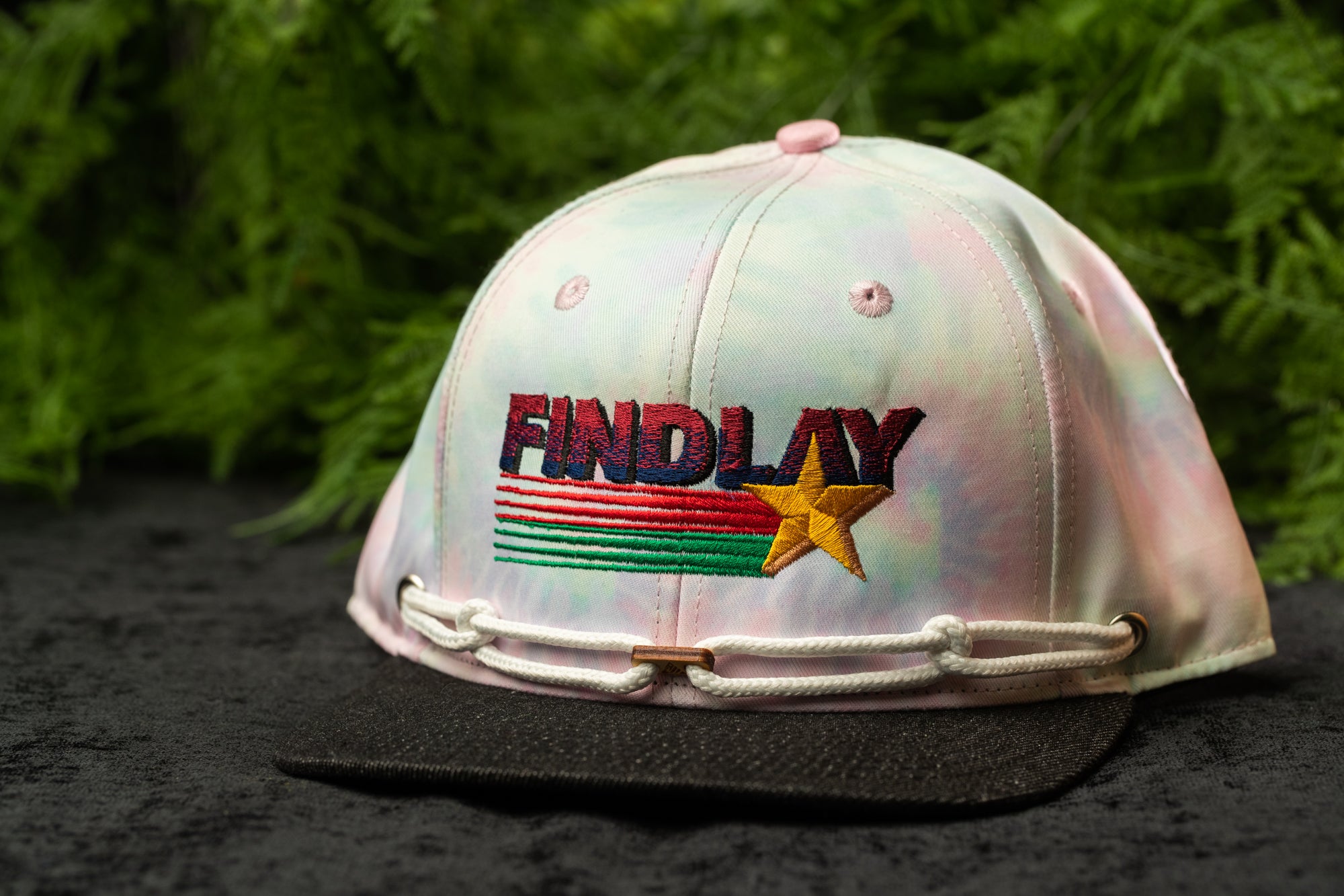A tie-dye baseball cap with the word "FINDA" printed on it, sitting on a black surface against a background of green foliage.