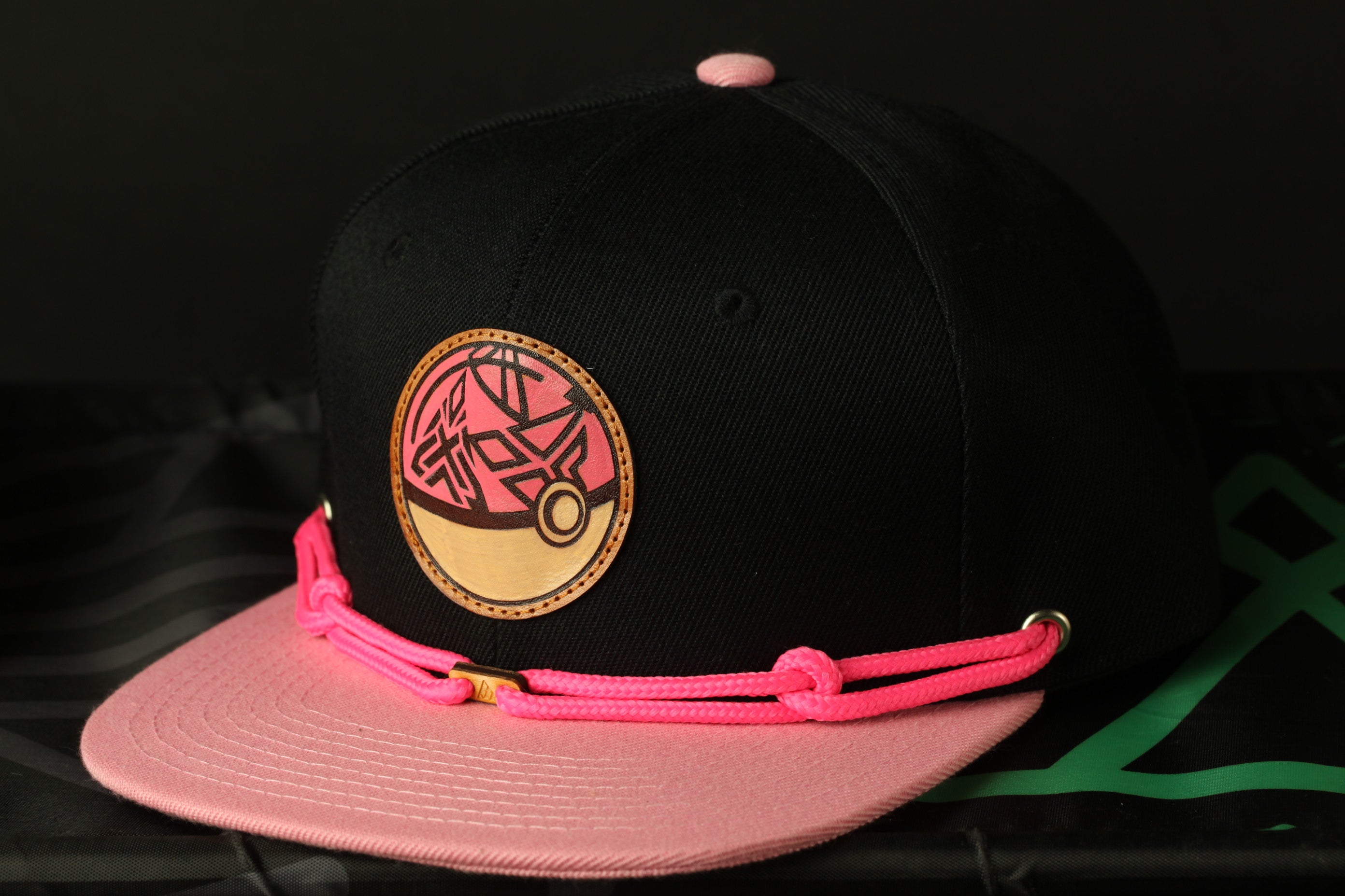 A black baseball cap with a pink and black design on the front, featuring a circular emblem with a pink and black pattern.