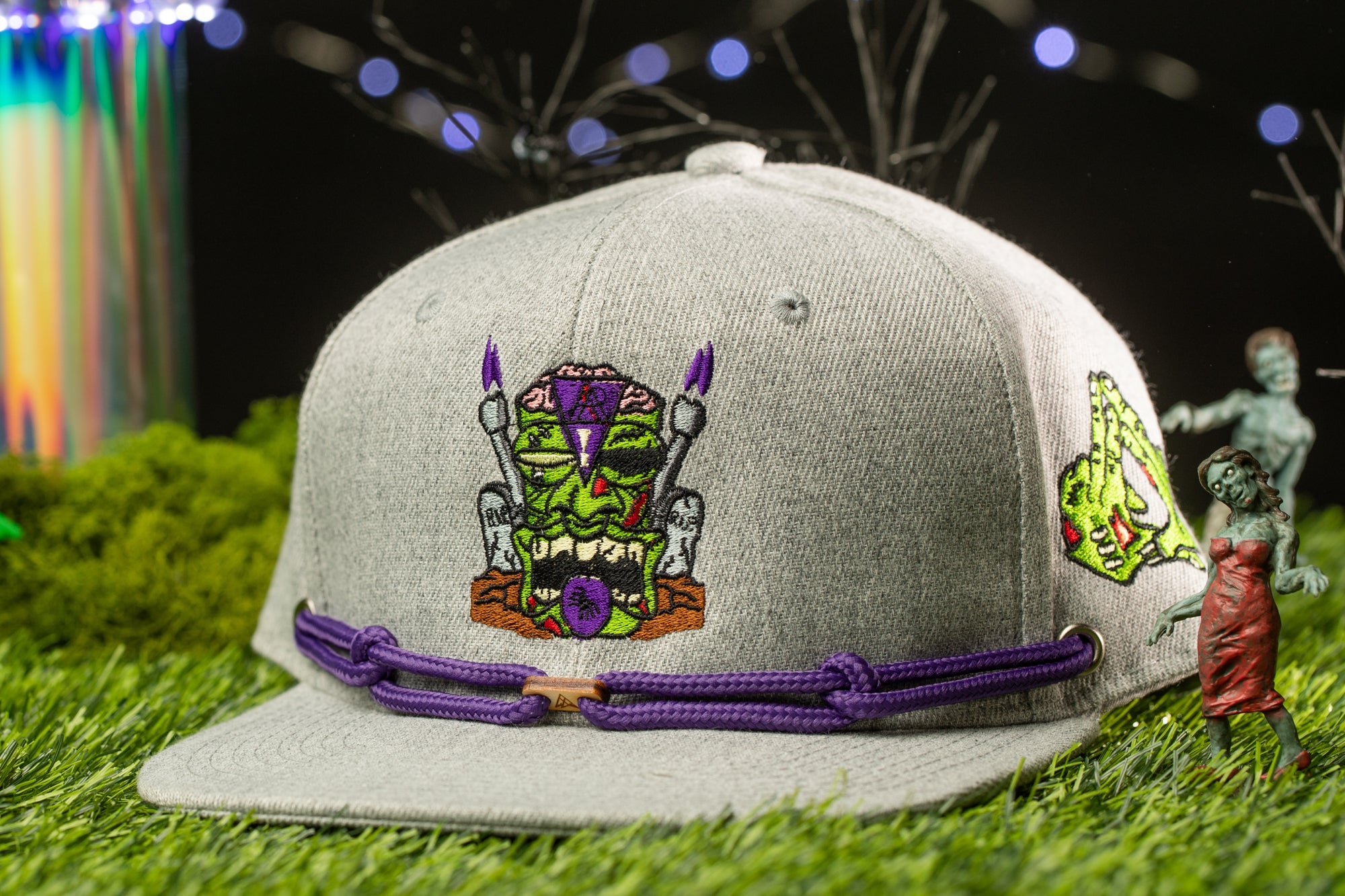 A gray baseball cap with a purple and green design on it, placed on a grassy surface with a small figurine of a zombie-like character standing next to it.