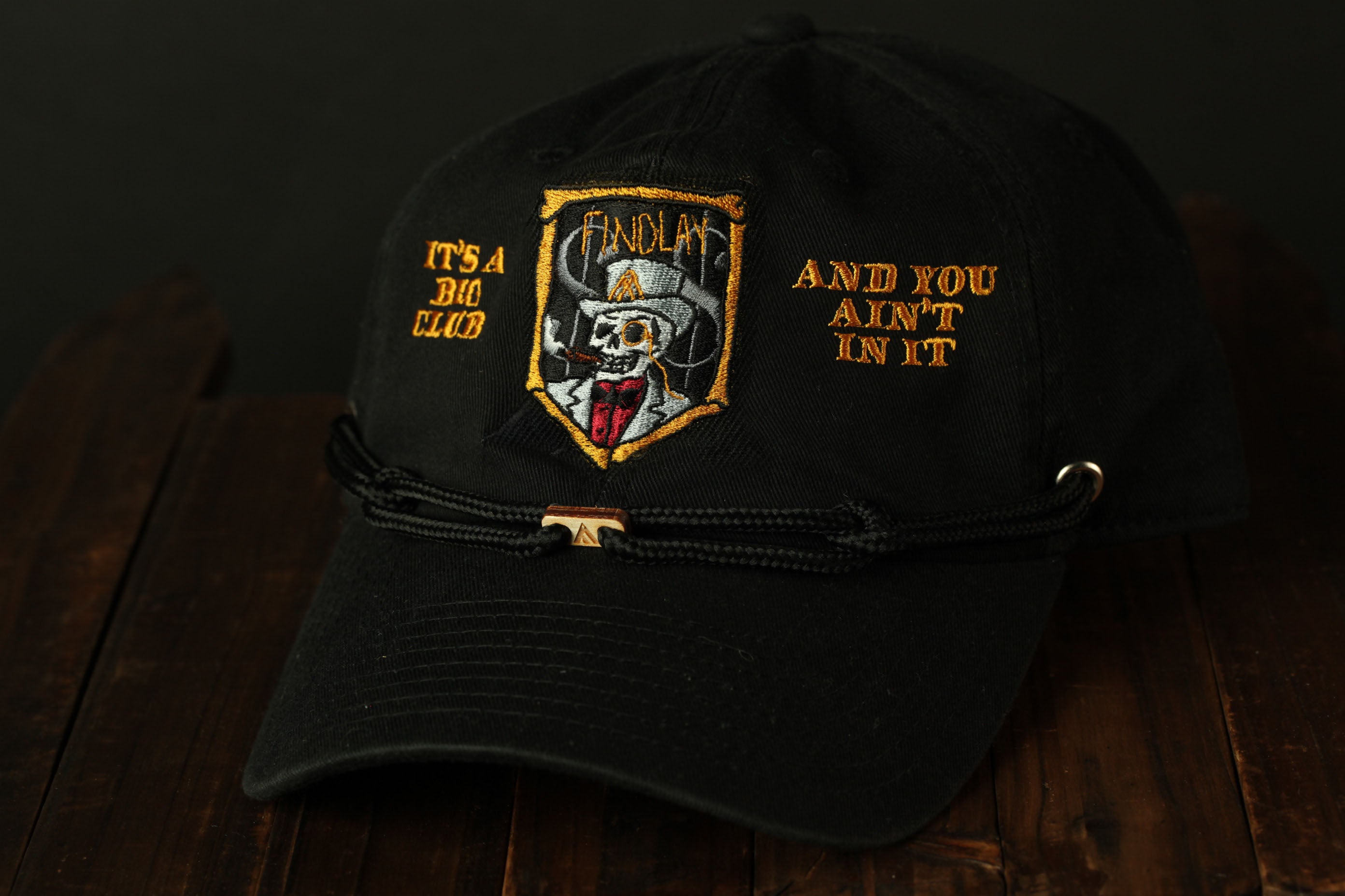 A black baseball cap with a skull and crossbones design, along with the text "IT'S A BIO CLUB AND YOU AIN'T IN IT" embroidered on it.