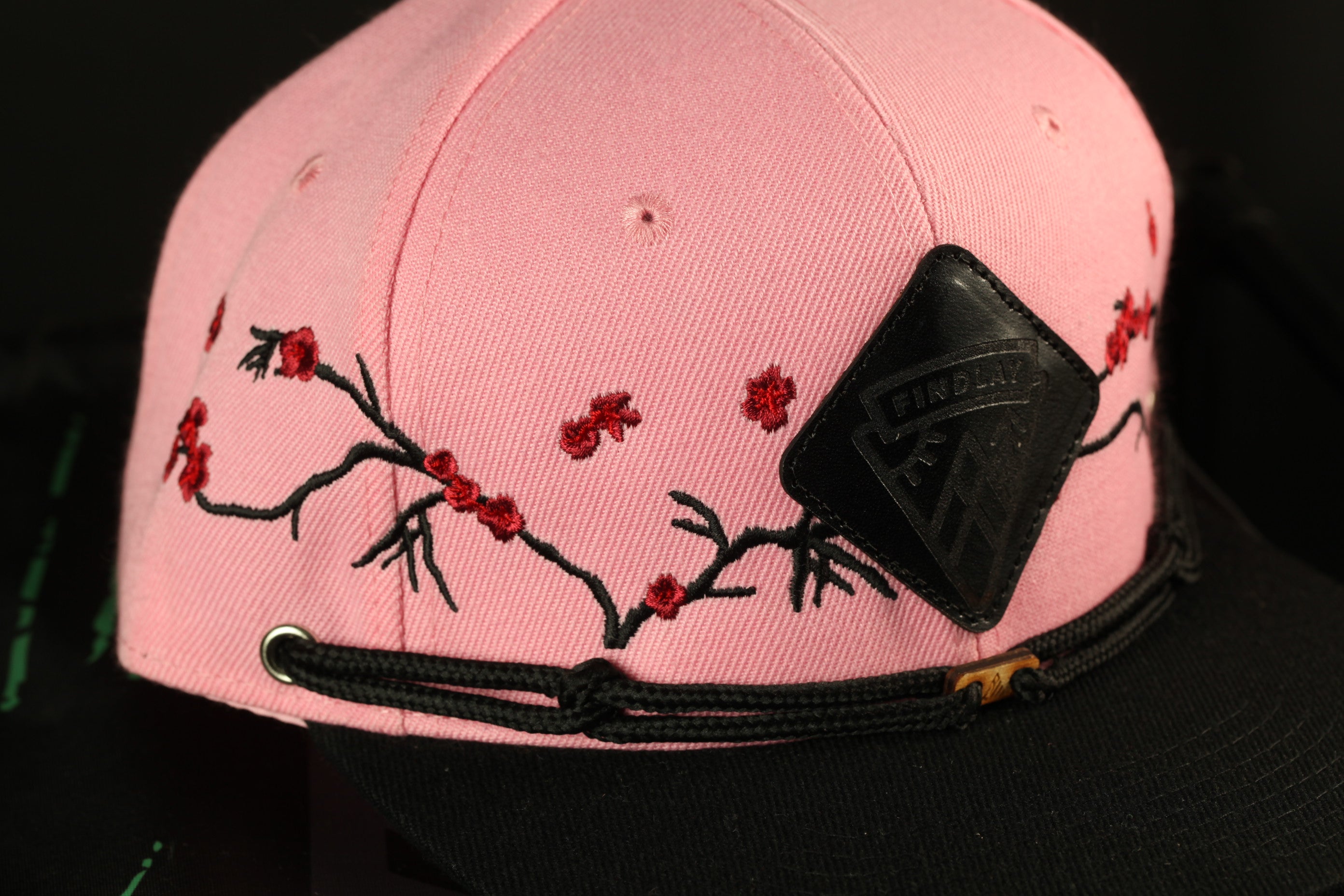A close-up view of a pink baseball cap with a black band and a black patch featuring red flowers on the brim.