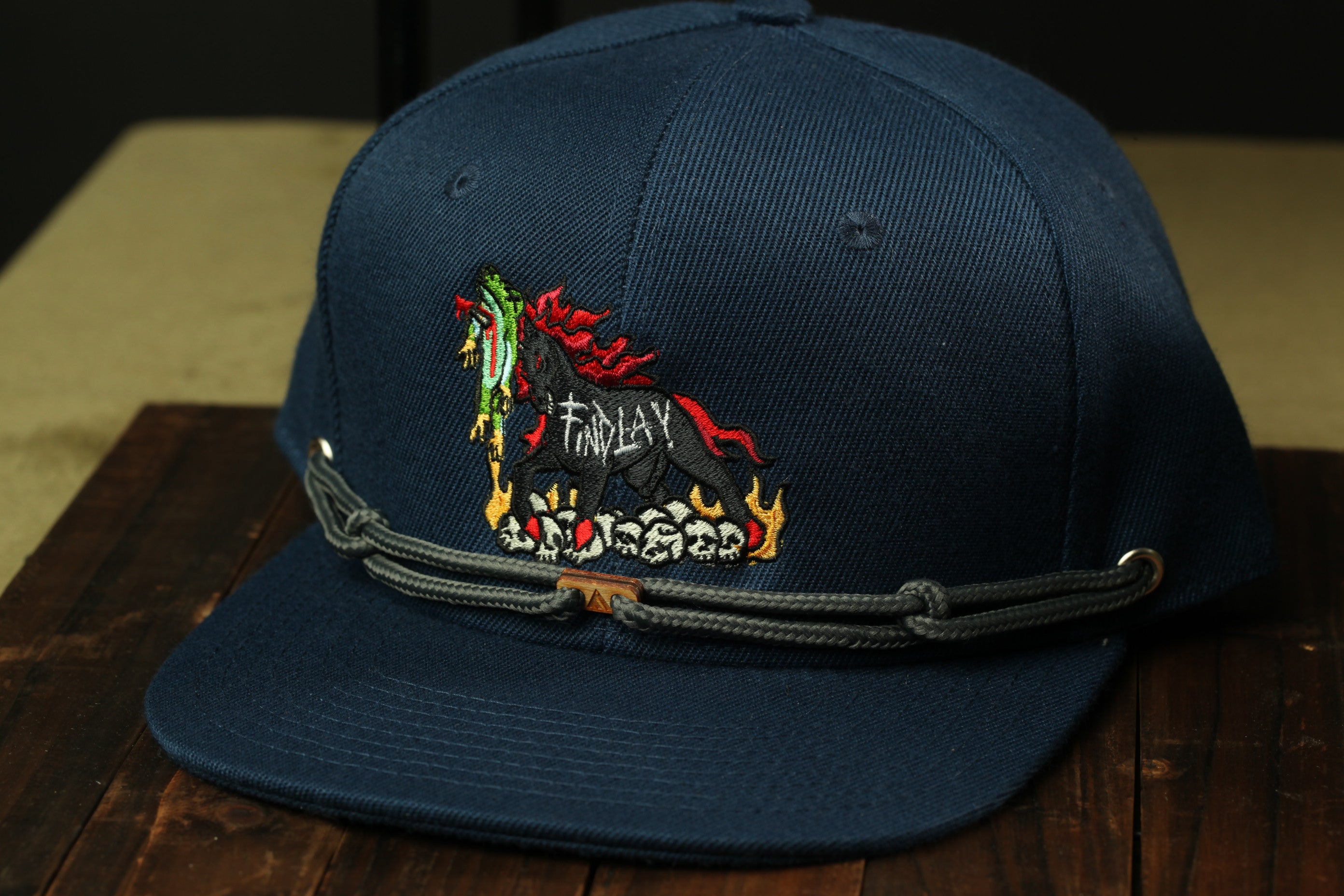 A navy blue baseball cap with a colorful embroidered design on the front, featuring a stylized animal or creature with flames or fire-like elements.