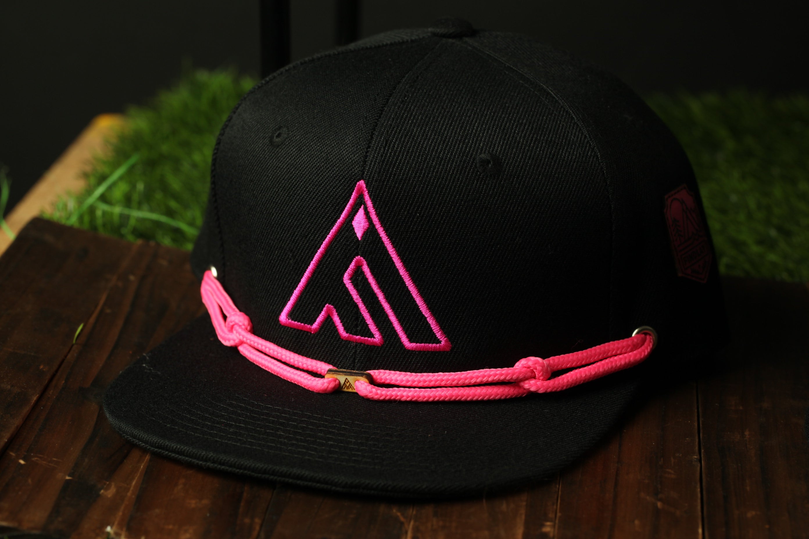 A black baseball cap with a pink logo on it, sitting on a wooden surface.