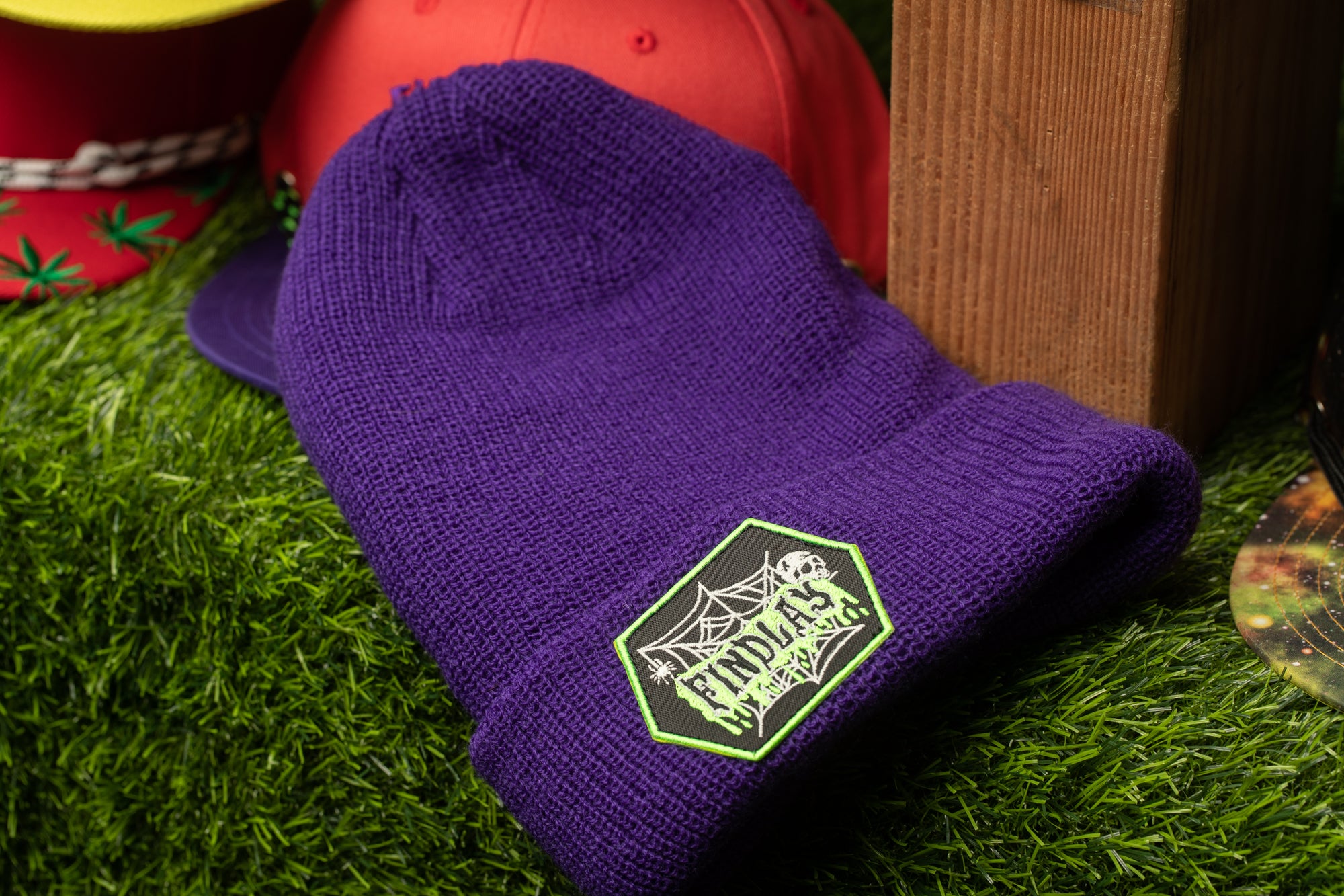 A purple beanie with a spider web design sits on a grassy surface, surrounded by other colorful items.