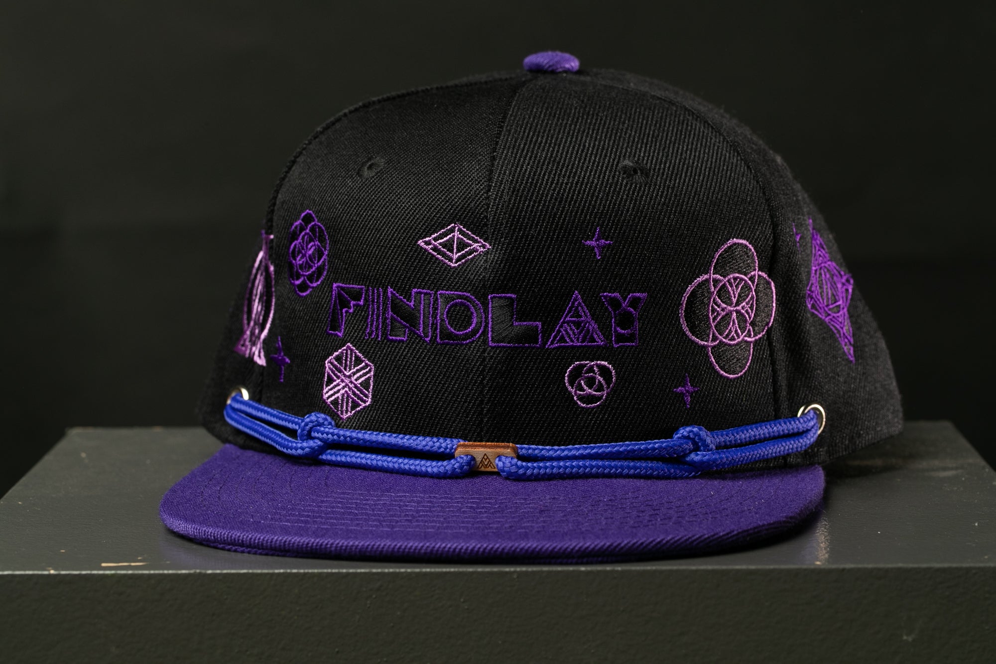 A black baseball cap with purple embroidered designs and the word "FINDLAY" on it.