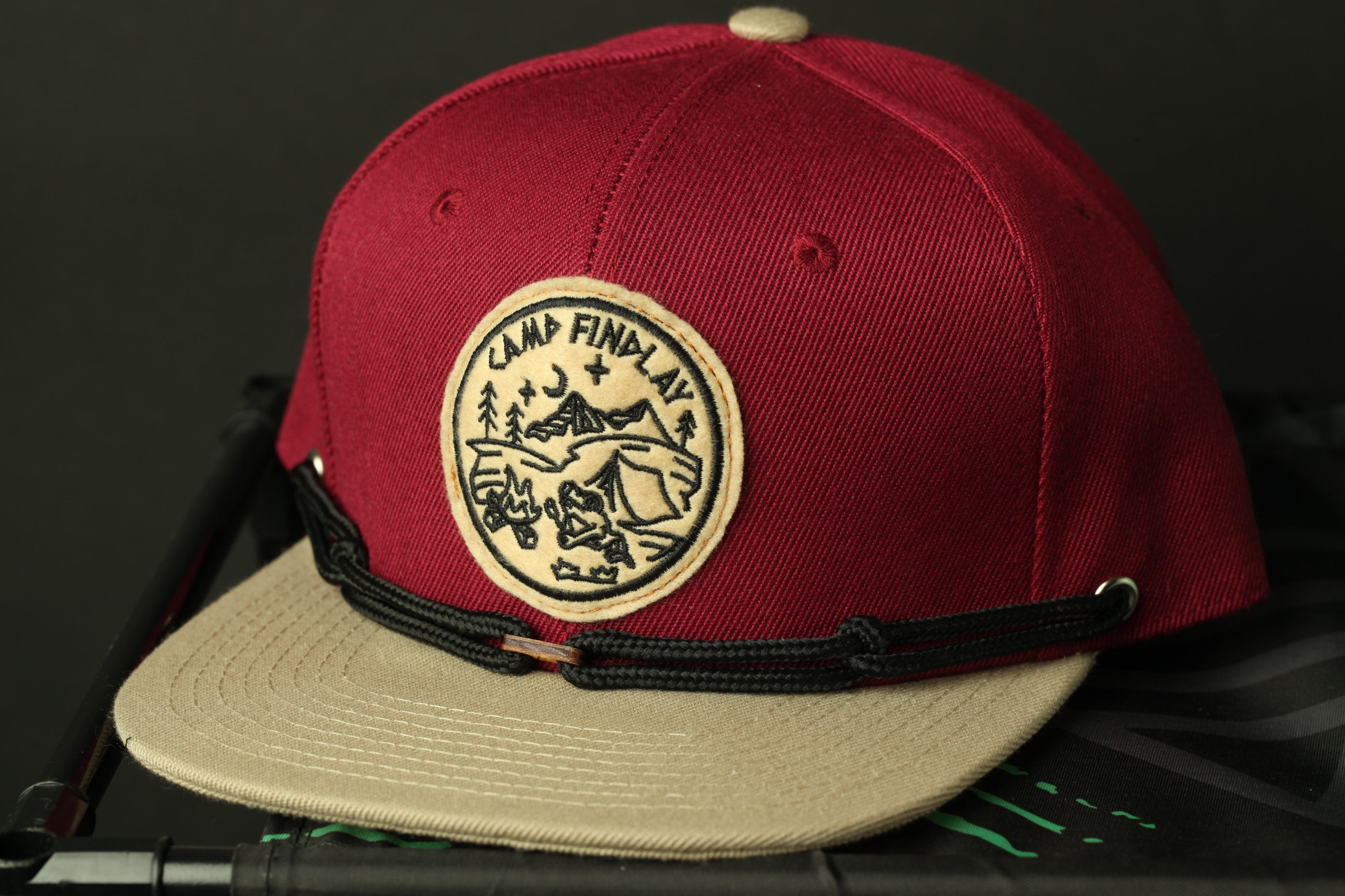 A red baseball cap with a circular patch on the front, featuring a mountain landscape and the text "CAMP FIND AL".