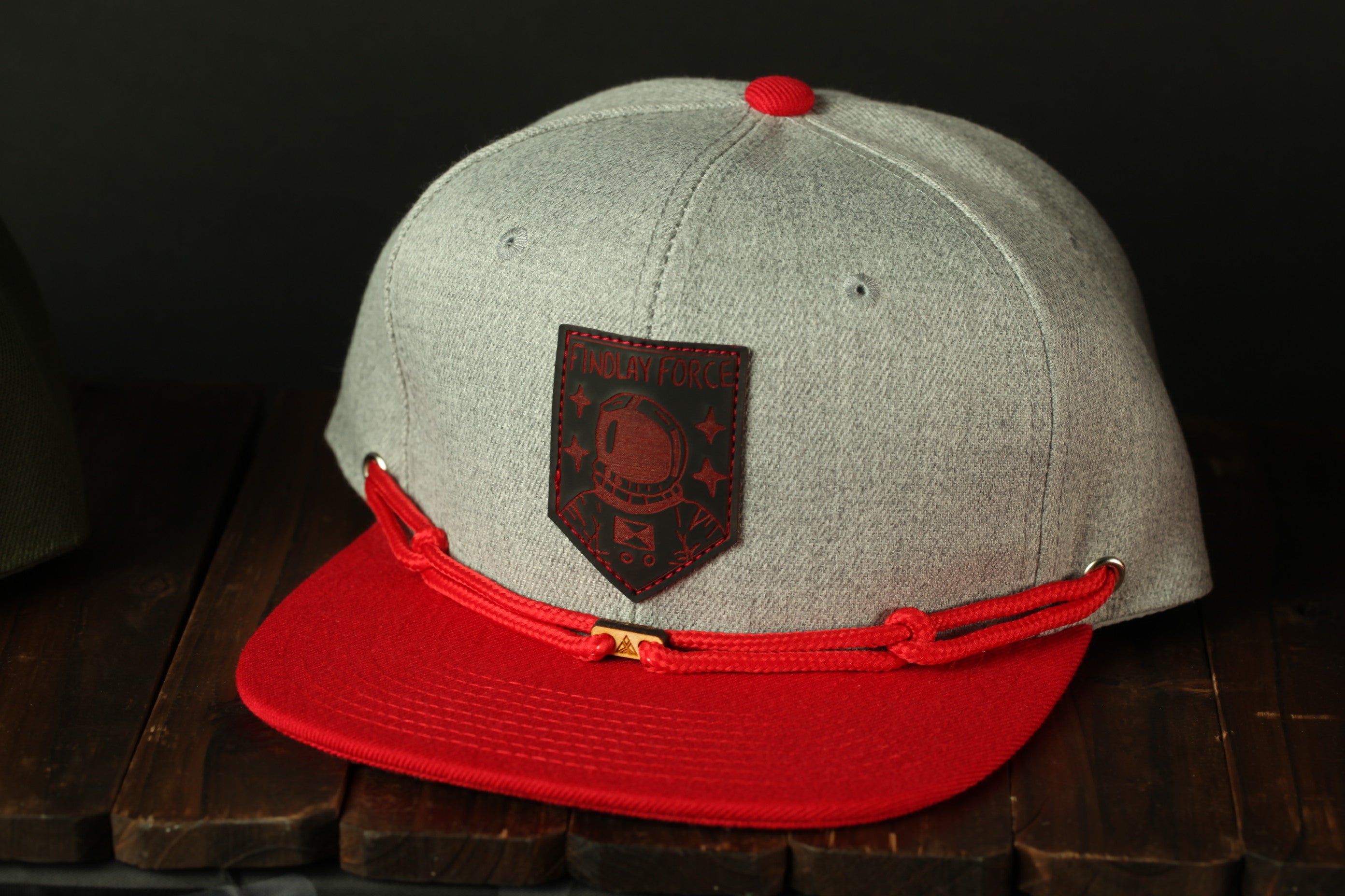 A gray baseball cap with a red brim and a black patch featuring a red helmet and the text "INDOLAY FORCE" is displayed on a wooden surface.