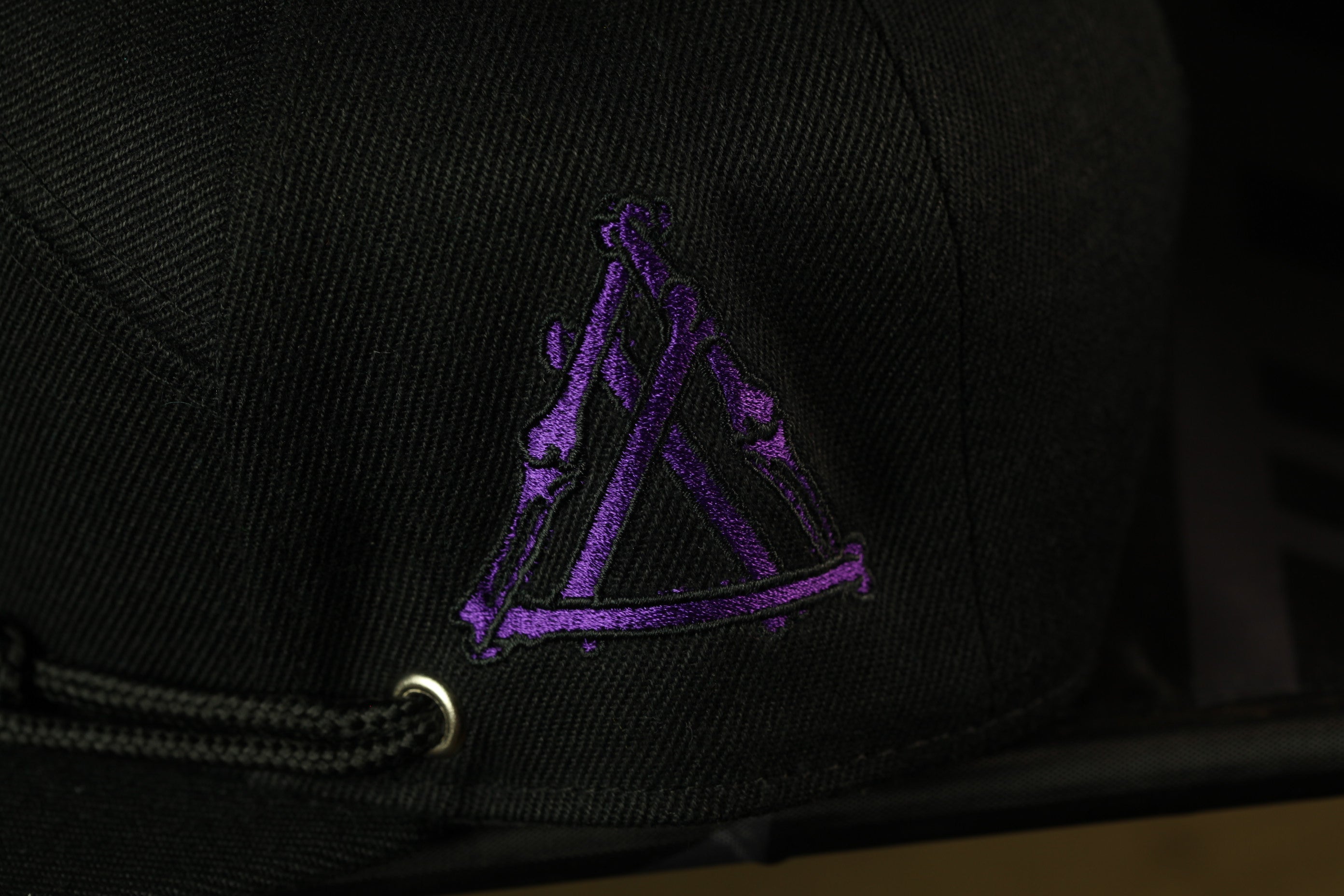 A close-up view of a black baseball cap with a purple embroidered logo on the front.