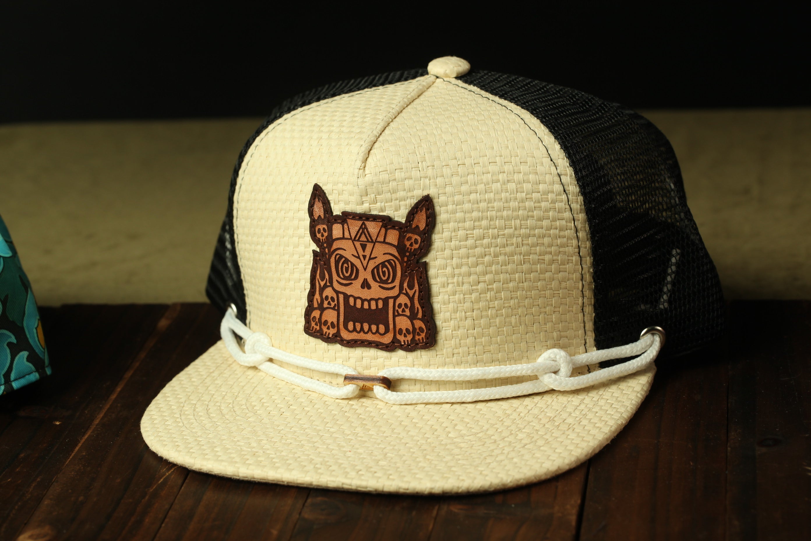 A beige baseball cap with a black mesh back and a white rope strap, featuring a cartoon-style skull design on the front.